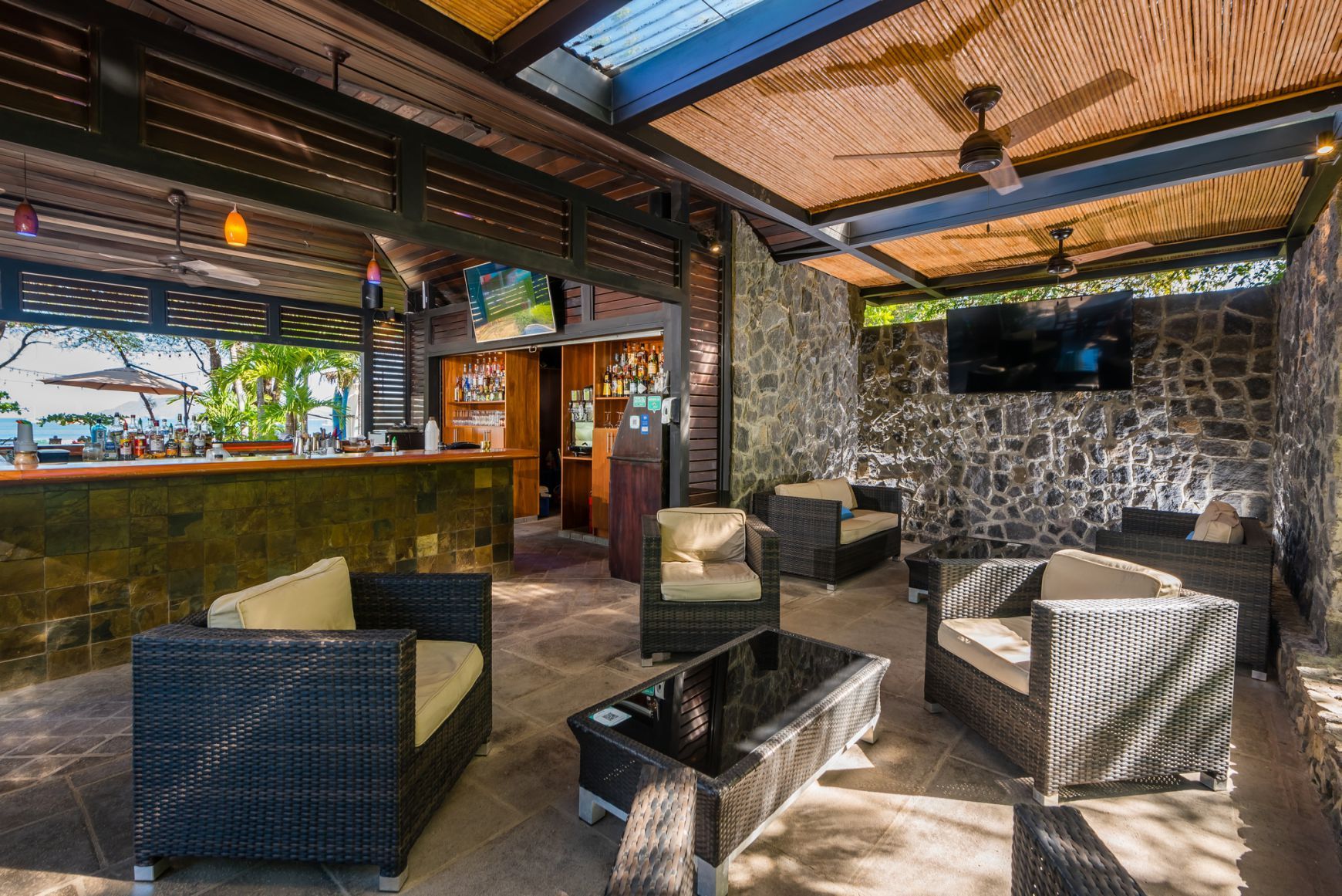 Outdoor lounge with wicker chairs and a stone wall under a covered patio with skylights and TV