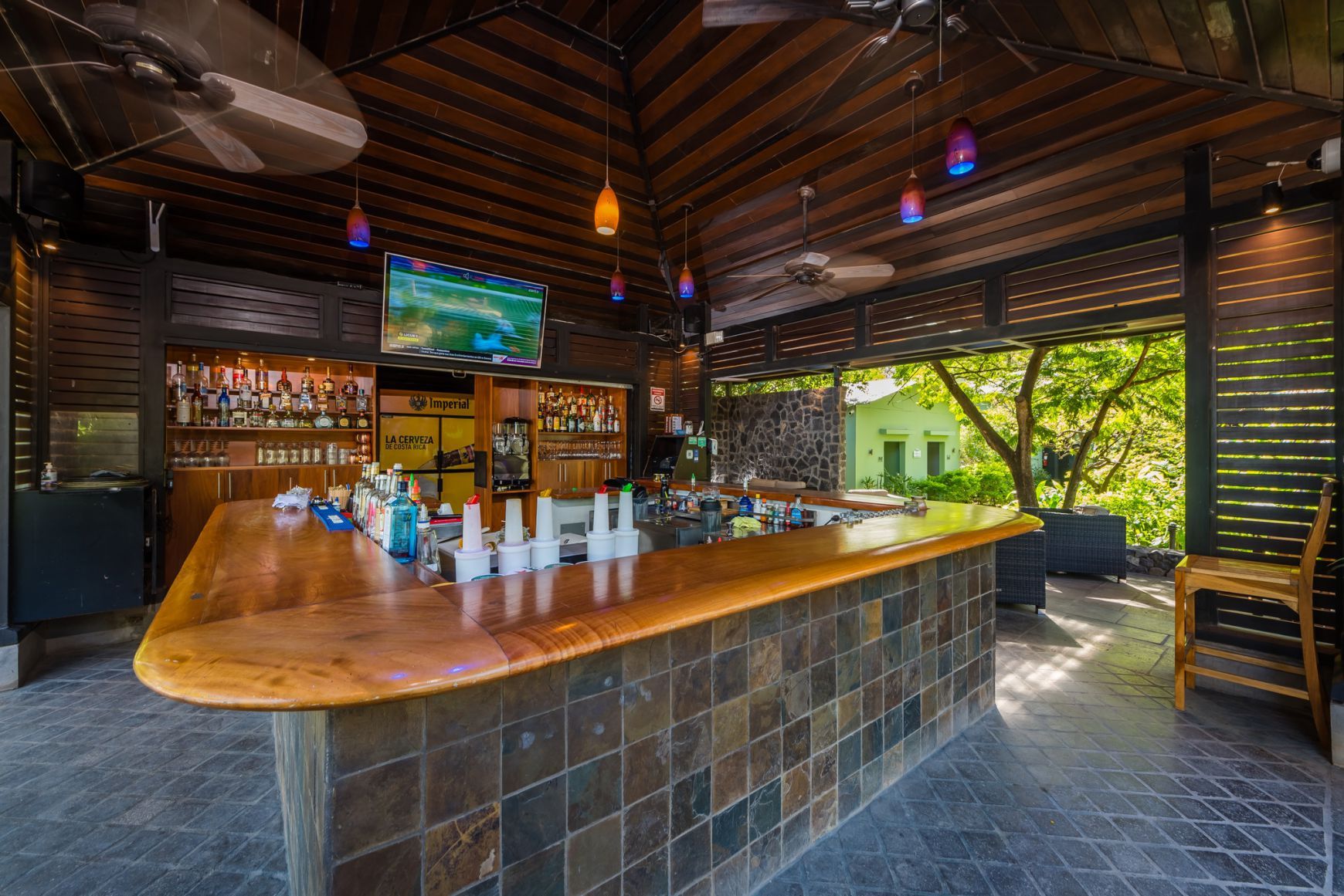 Rustic outdoor bar with wooden counter, hanging lights, and bottles under a thatched roof.