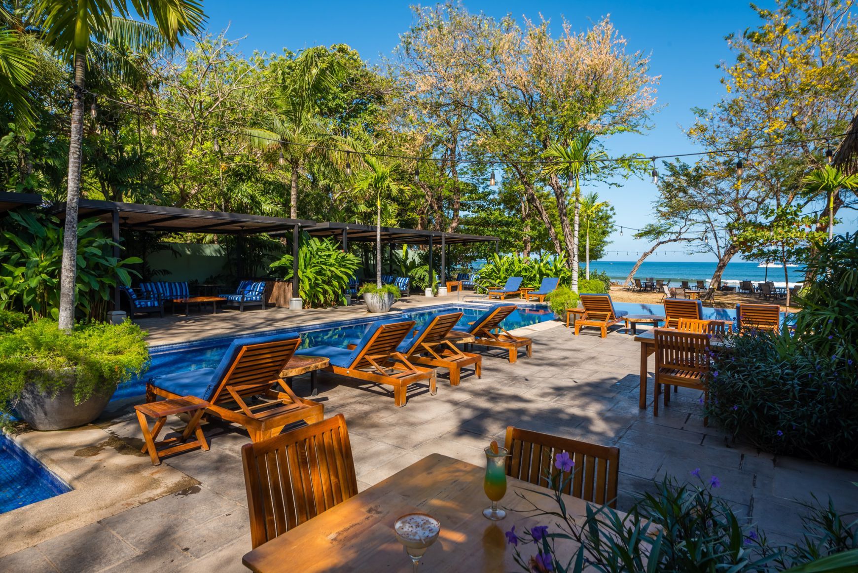 Beachfront café patio with wooden lounge chairs, tables, umbrellas, and ocean view under palm trees