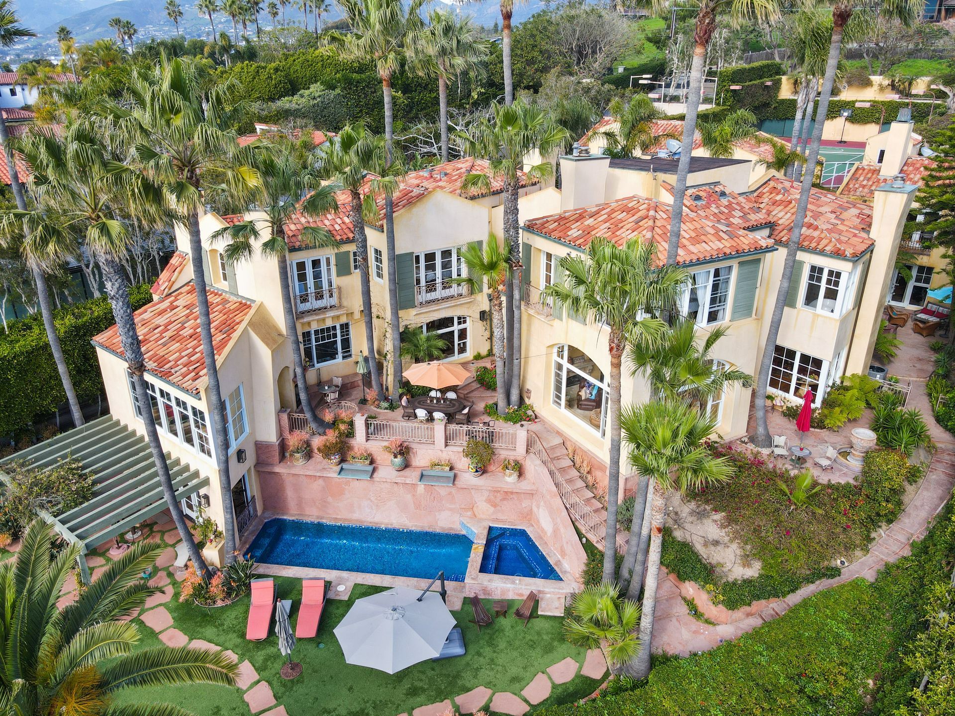 Aerial view of a tropical villa with red tile roofs, a pool, patio loungers, and palm trees.