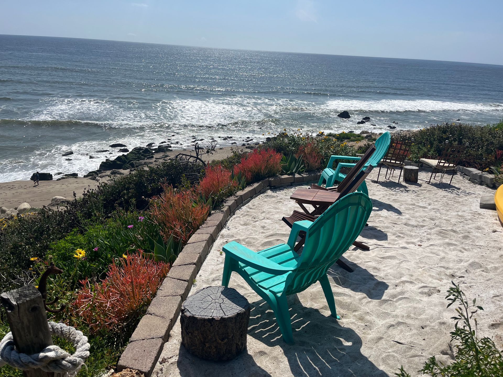 Turquoise chairs on a sandy patio overlooking a rocky beach and sparkling ocean