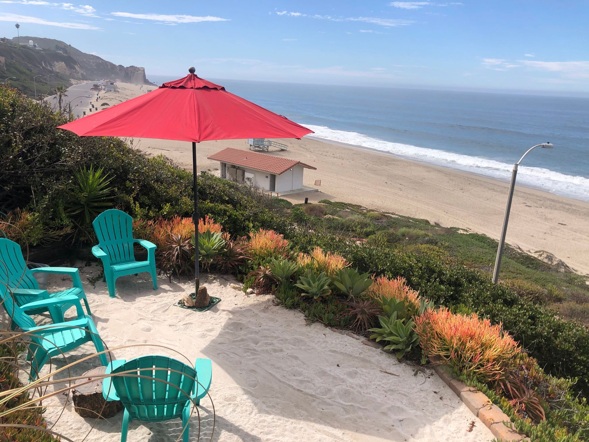 Beach view with turquoise chairs and a red umbrella on a sandy patio above the ocean