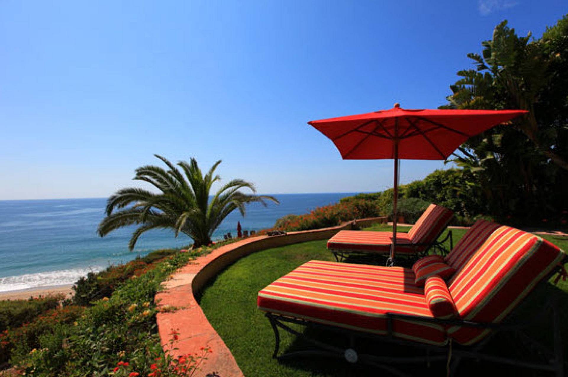 Oceanfront patio with red lounge chairs and umbrella overlooking a palm tree and blue sea