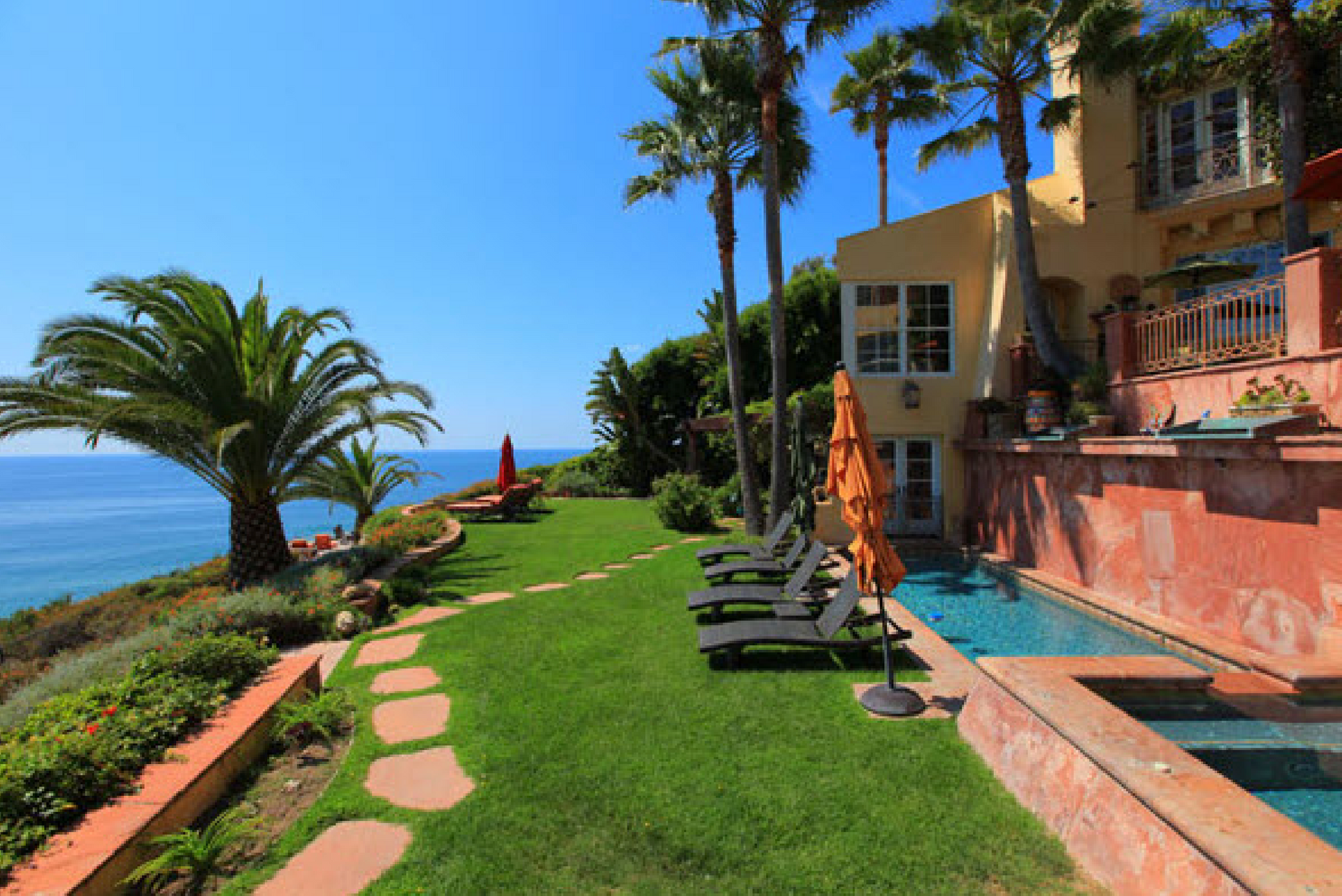 Oceanfront resort patio with lounge chairs, green lawn, palm trees, and clear blue sky.