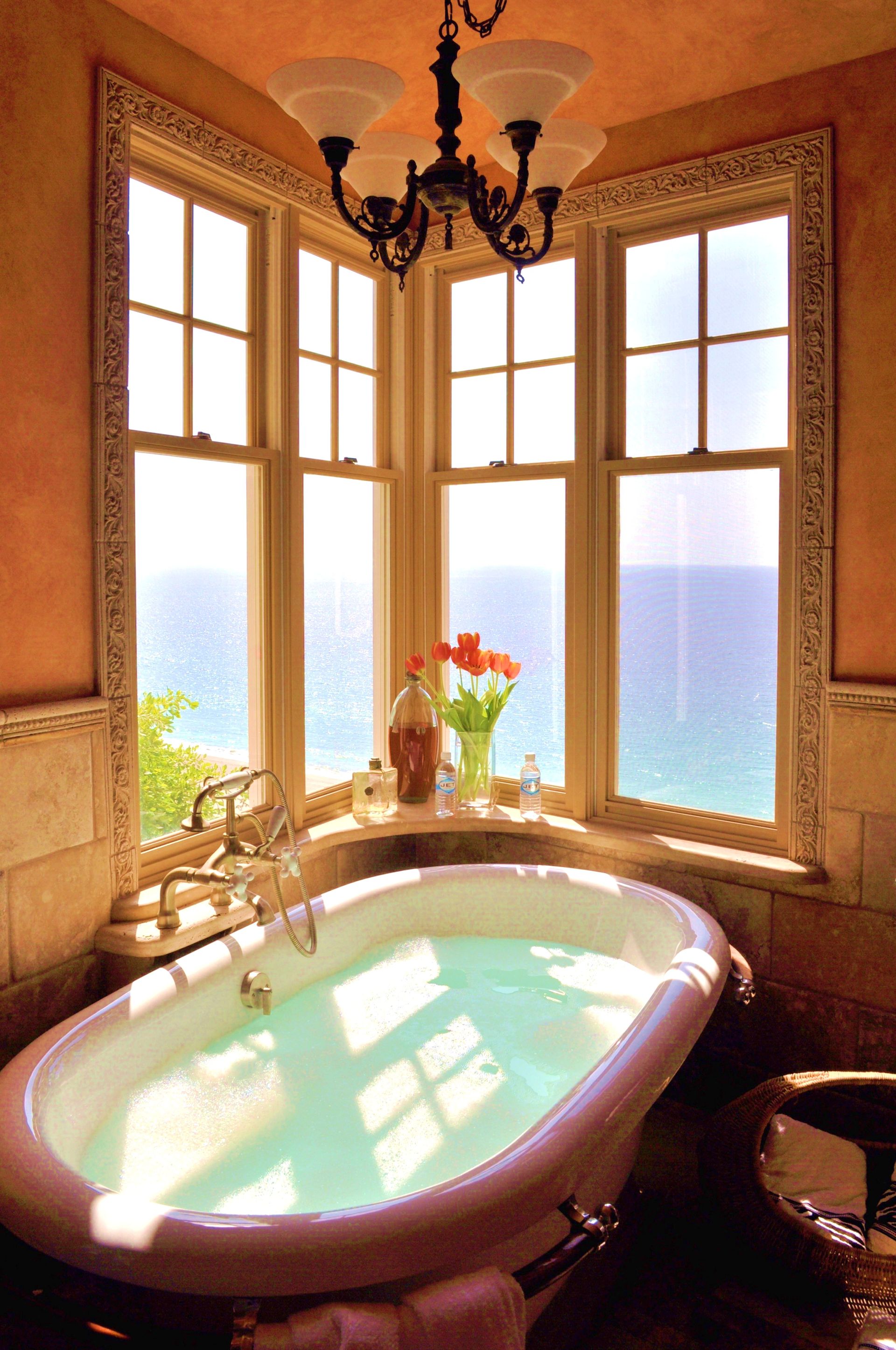 Sunlit bathroom with a clawfoot tub beside tall windows overlooking the ocean