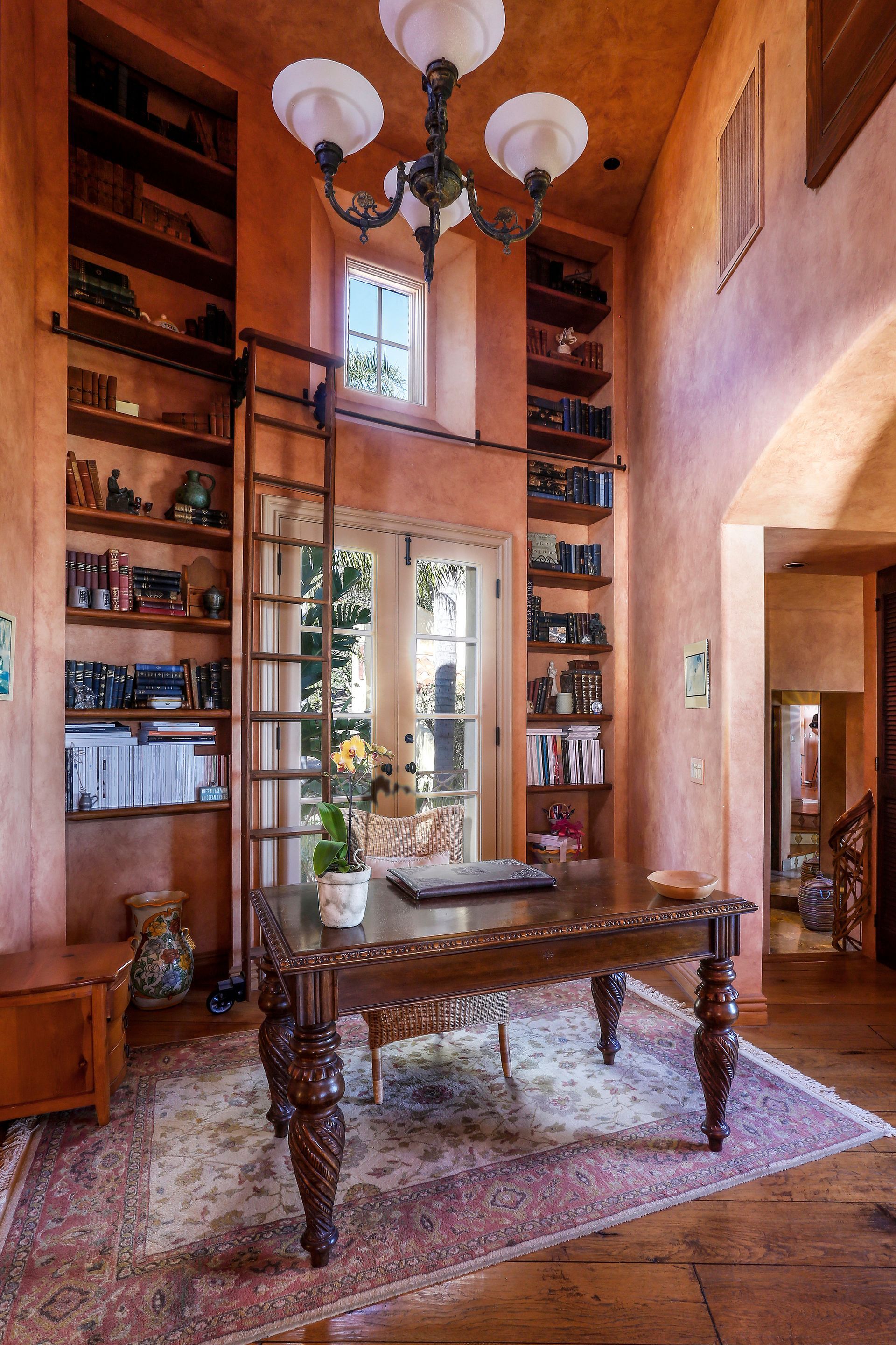 Tall foyer with wood shelves, ornate table, and chandelier, lit by a central glass door and window.