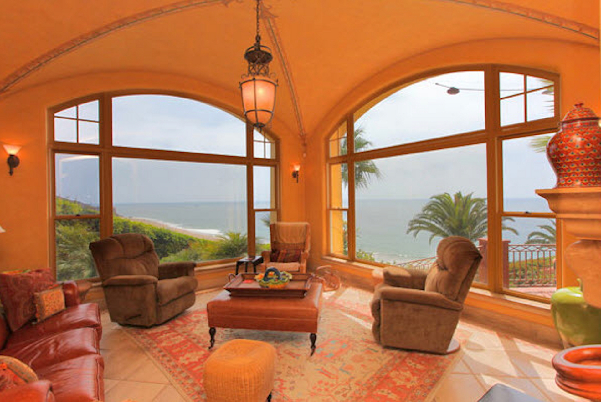 Warm living room with arched windows, ocean view, leather chairs, sofa, and lamp on a patterned rug