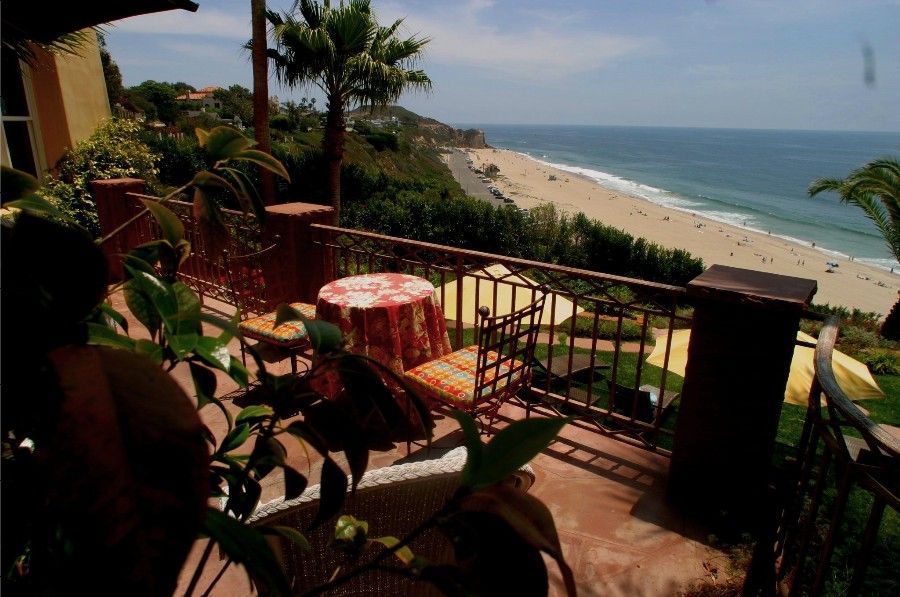 Balcony patio overlooking a sandy beach and ocean, with a table, chairs, and potted plants in sunlight