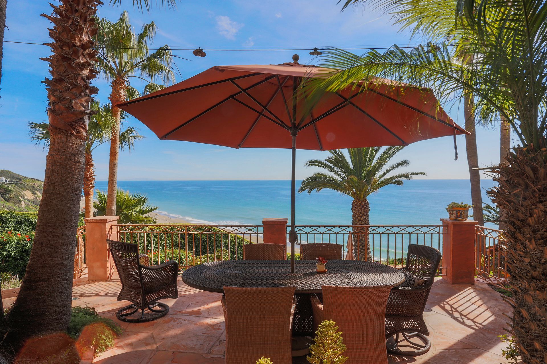 Patio with red umbrella, wicker chairs, and ocean view under palm trees