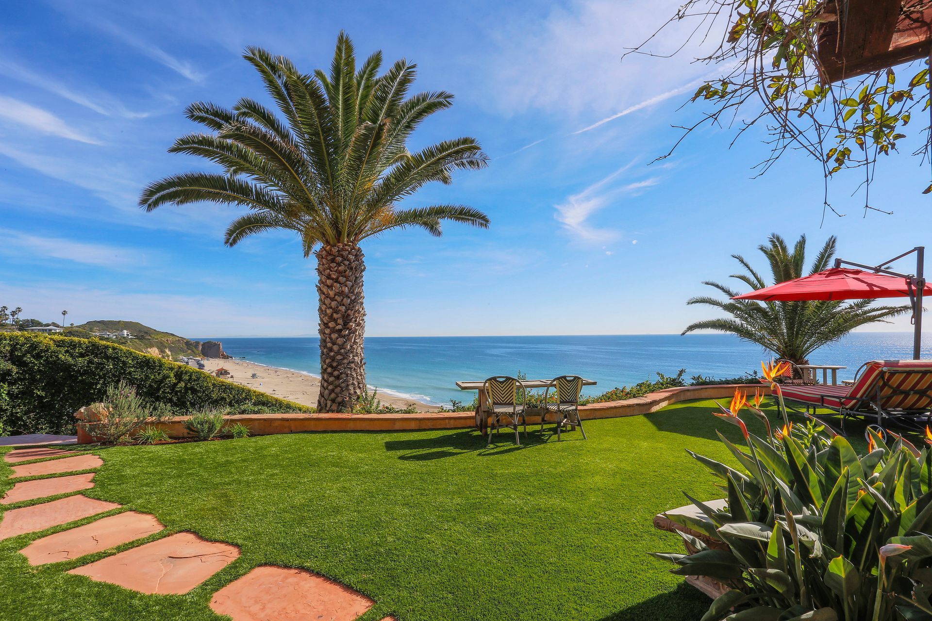 Tropical garden patio with palm tree, ocean view, stepping stones, and red umbrella seating