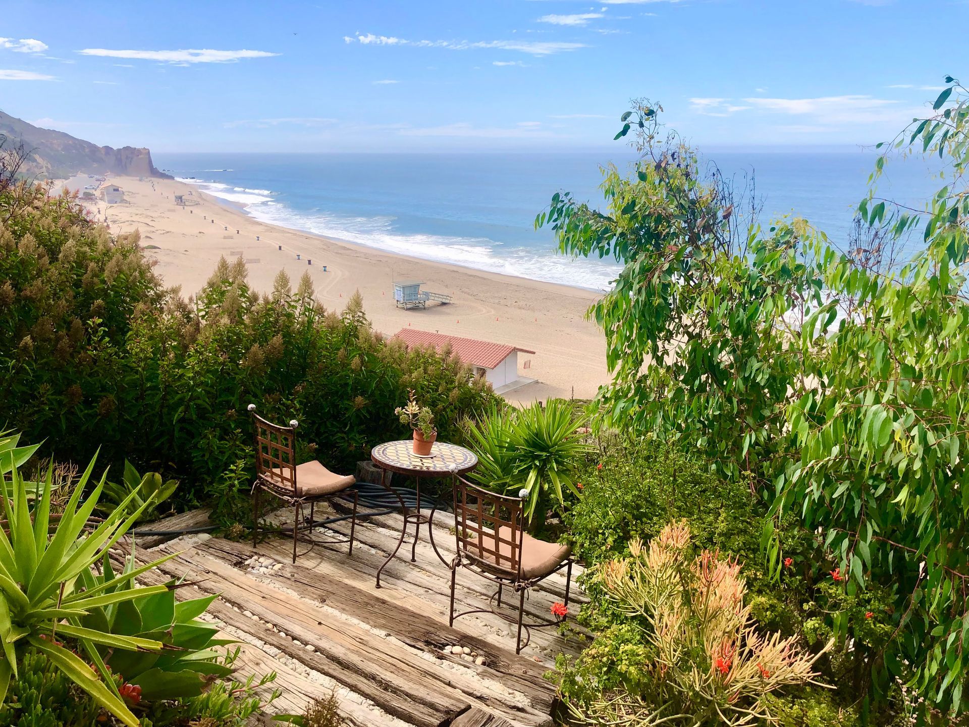 Sunny beach view from a terrace with chairs, table, and lush greenery overlooking the ocean