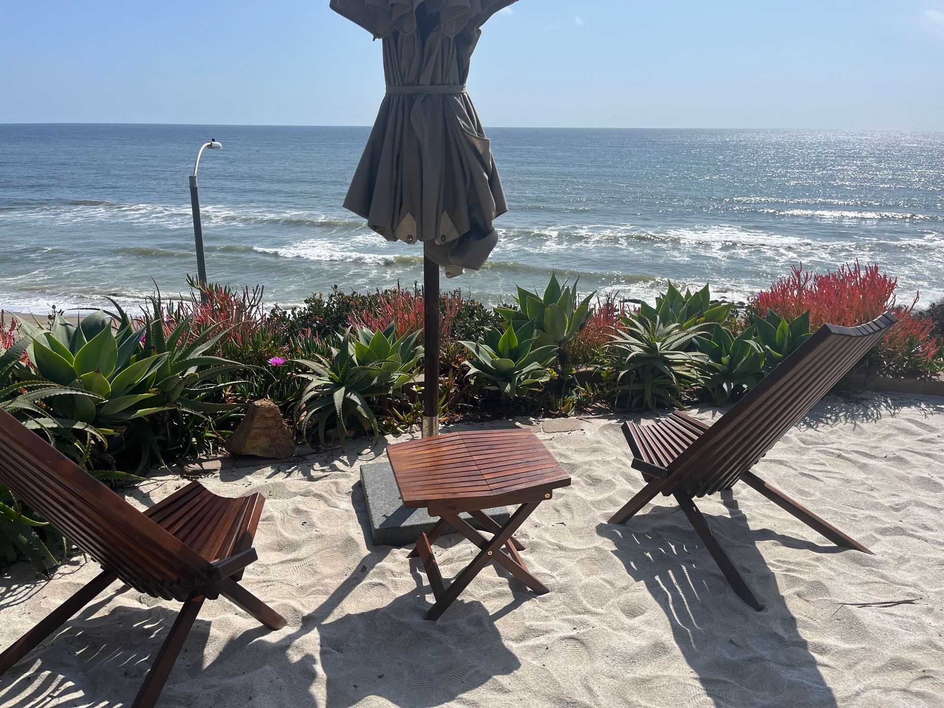 Beach patio with wooden lounge chairs, a closed umbrella, and ocean waves beyond flower beds