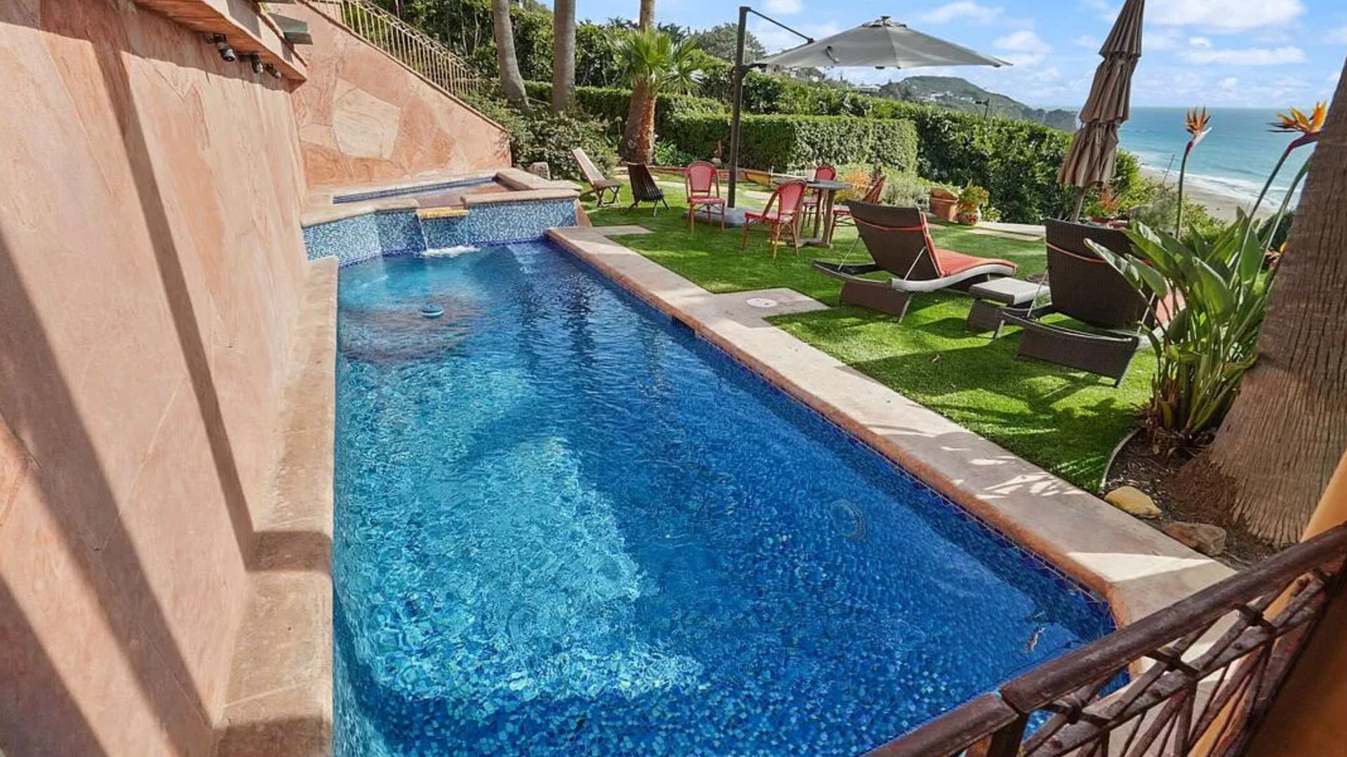 Long blue resort pool beside palm trees, lounge chairs, and ocean view on a sunny patio