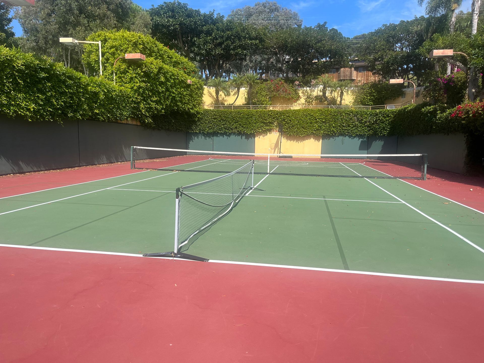 Empty outdoor tennis court with a green surface, red edges, and nets under a sunny sky.