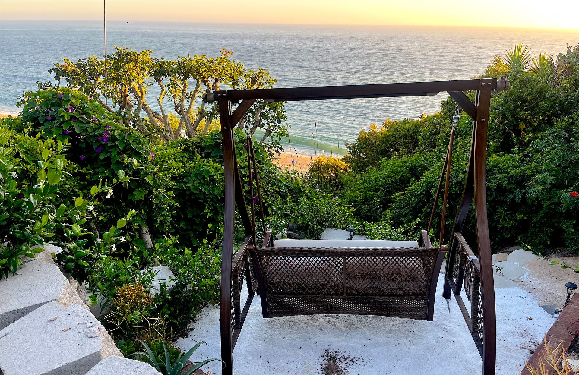 Wooden swing on a seaside terrace at sunset, framed by greenery and white steps