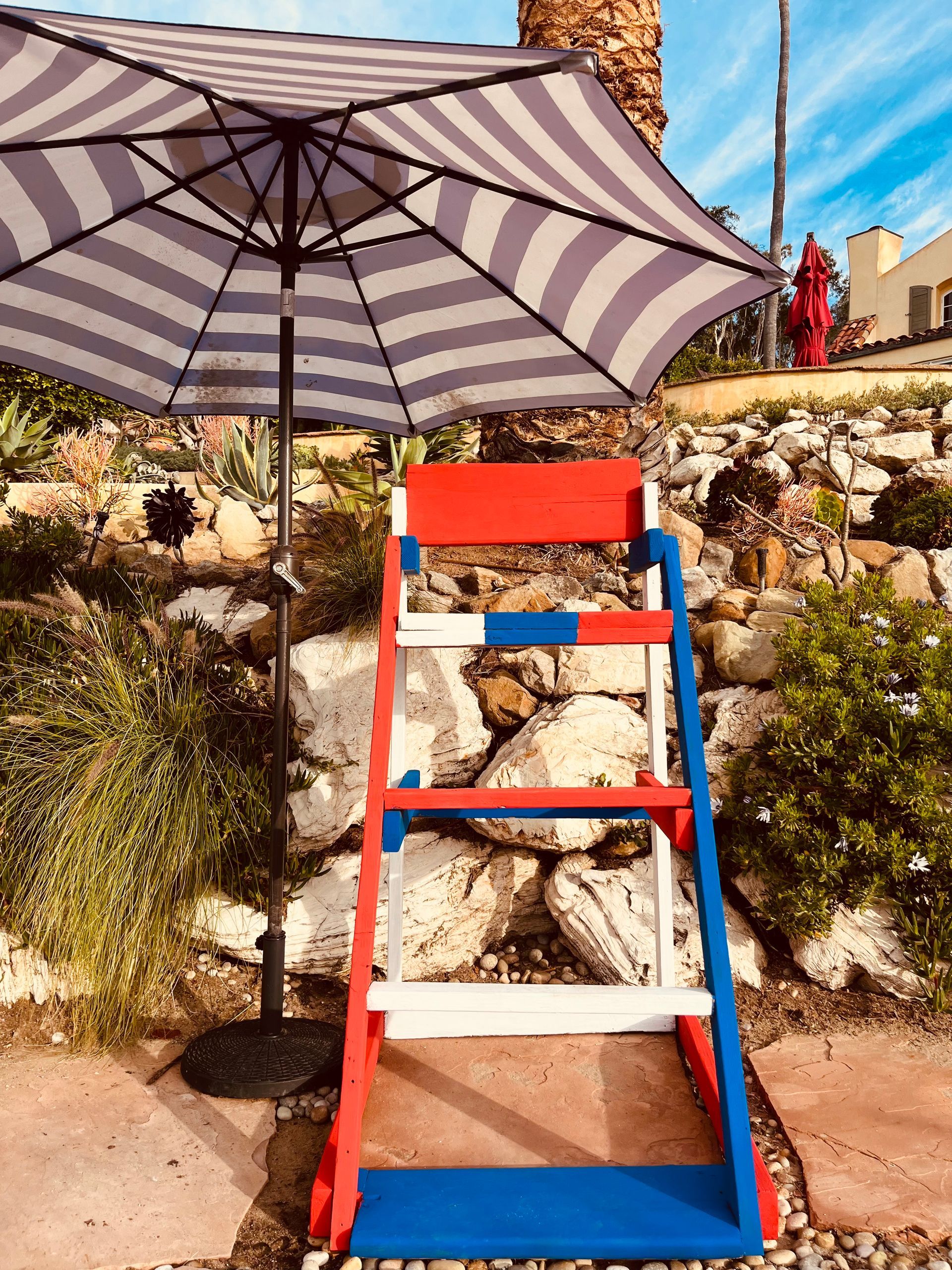 Red-and-blue lifeguard chair under a striped umbrella on a sunny outdoor terrace