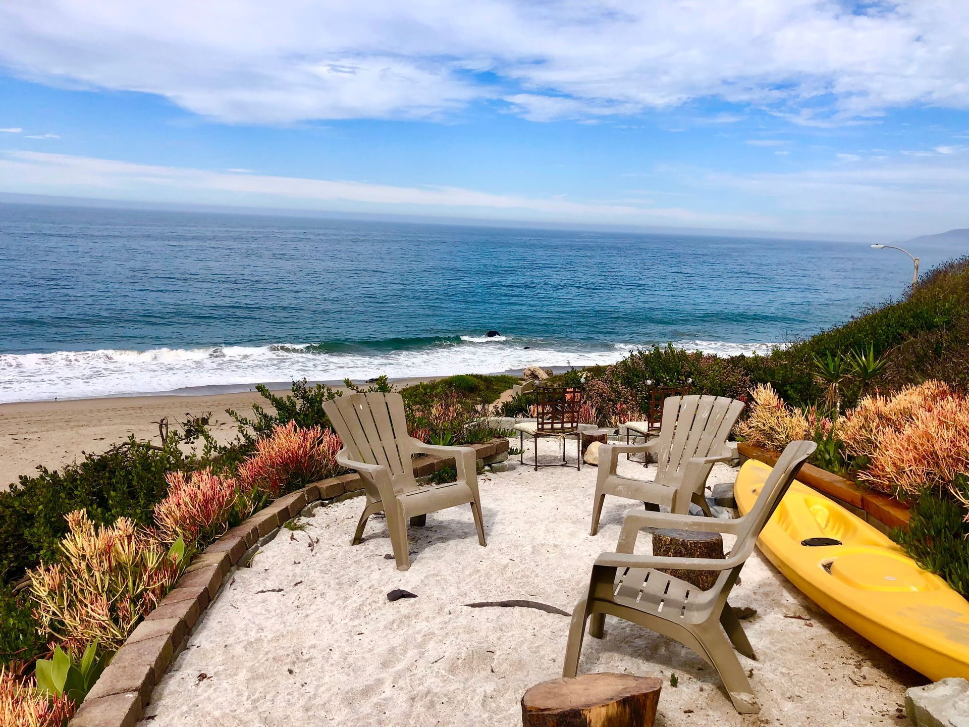 Patio chairs overlooking a calm ocean, with flowers and a yellow kayak by the shore.