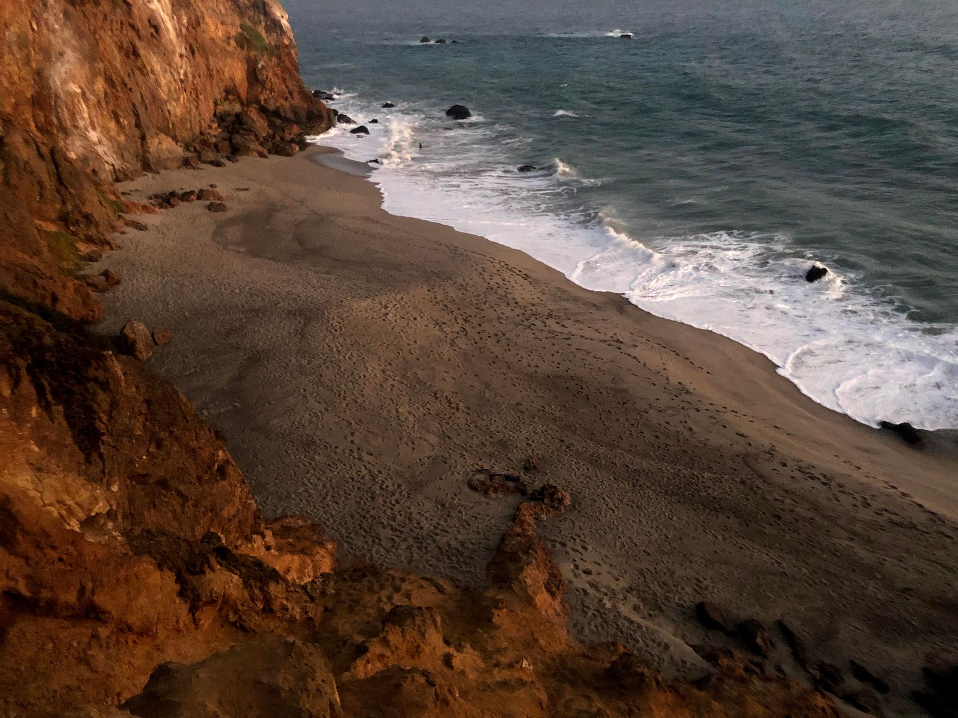 Rocky coastal cliff overlooking a gray sandy beach with waves crashing under an overcast sky