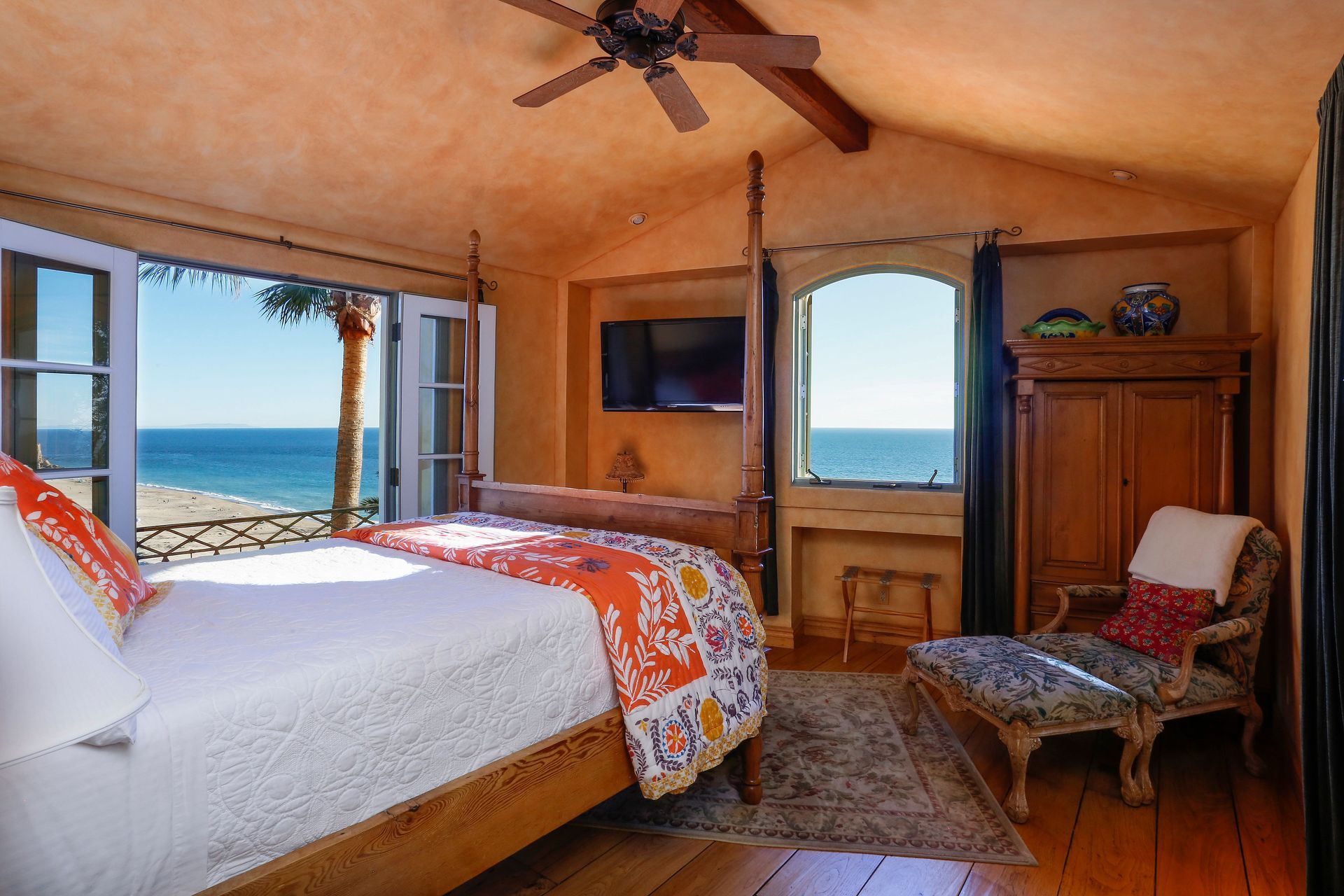 Cozy bedroom with ocean-view windows, a bed with orange bedding, and a wooden chair by the window.