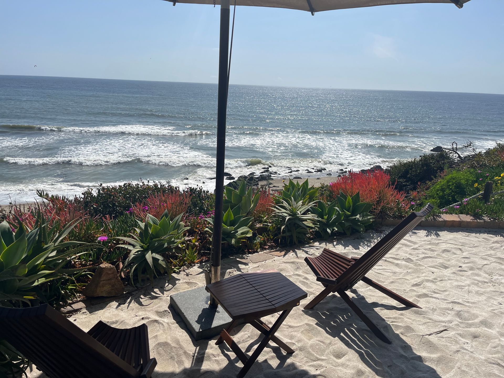 Beachside chairs and table under an umbrella overlooking a sunny ocean with waves.