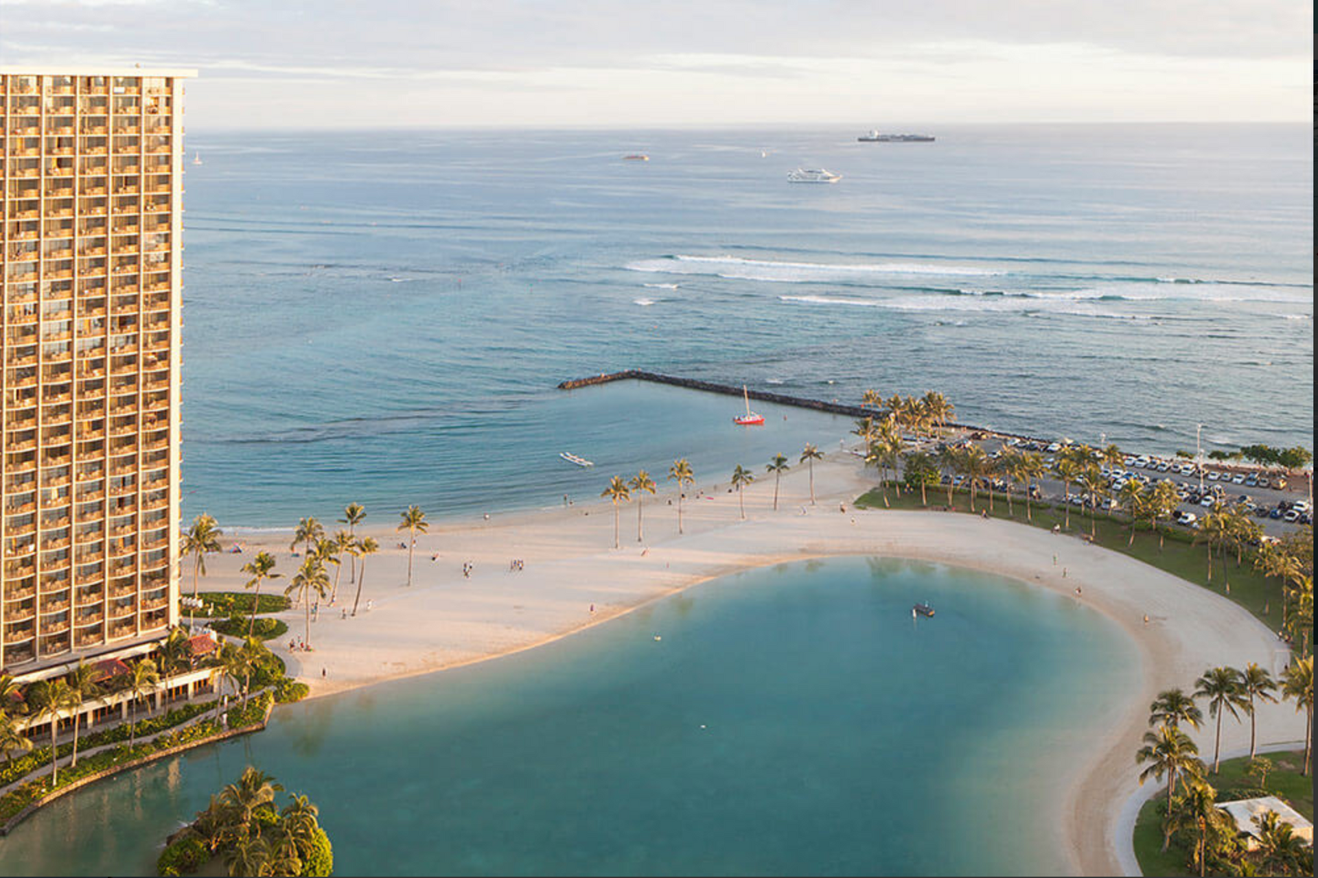 Aerial view of a beach resort with a turquoise lagoon, palm trees, and ocean waves beyond a tall building