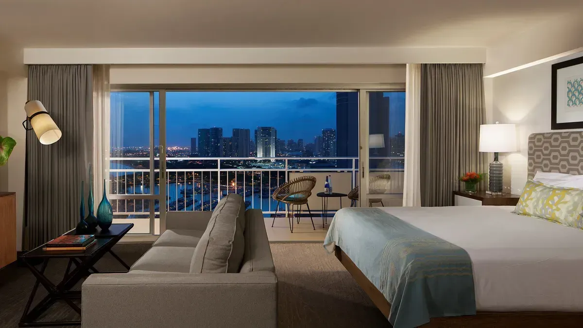 Modern hotel room with two beds, sofa, TV, and large balcony window overlooking a city skyline at dusk