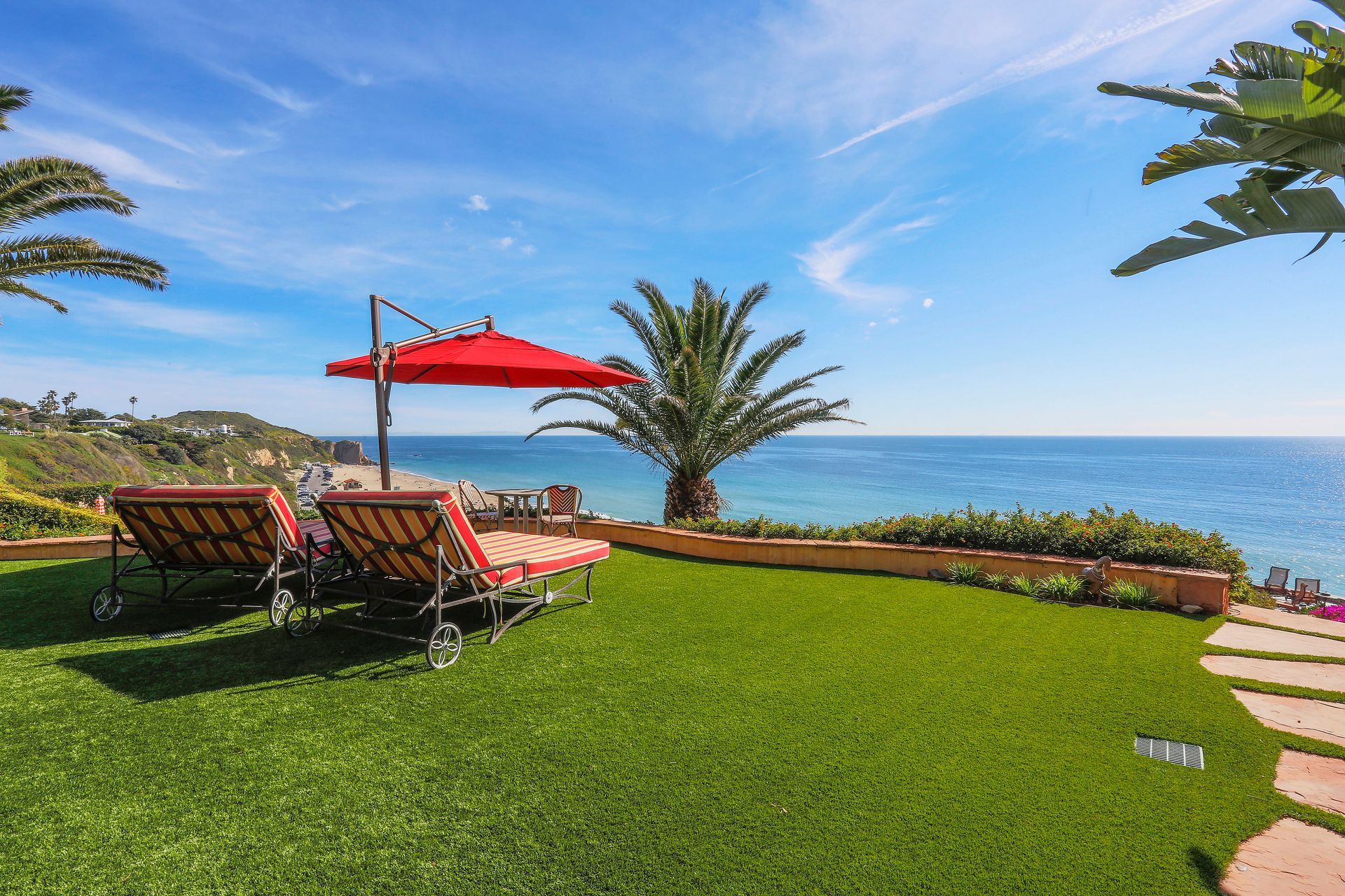 Oceanfront lawn with lounge chairs, red umbrella, palm tree, and clear blue sea under a sunny sky