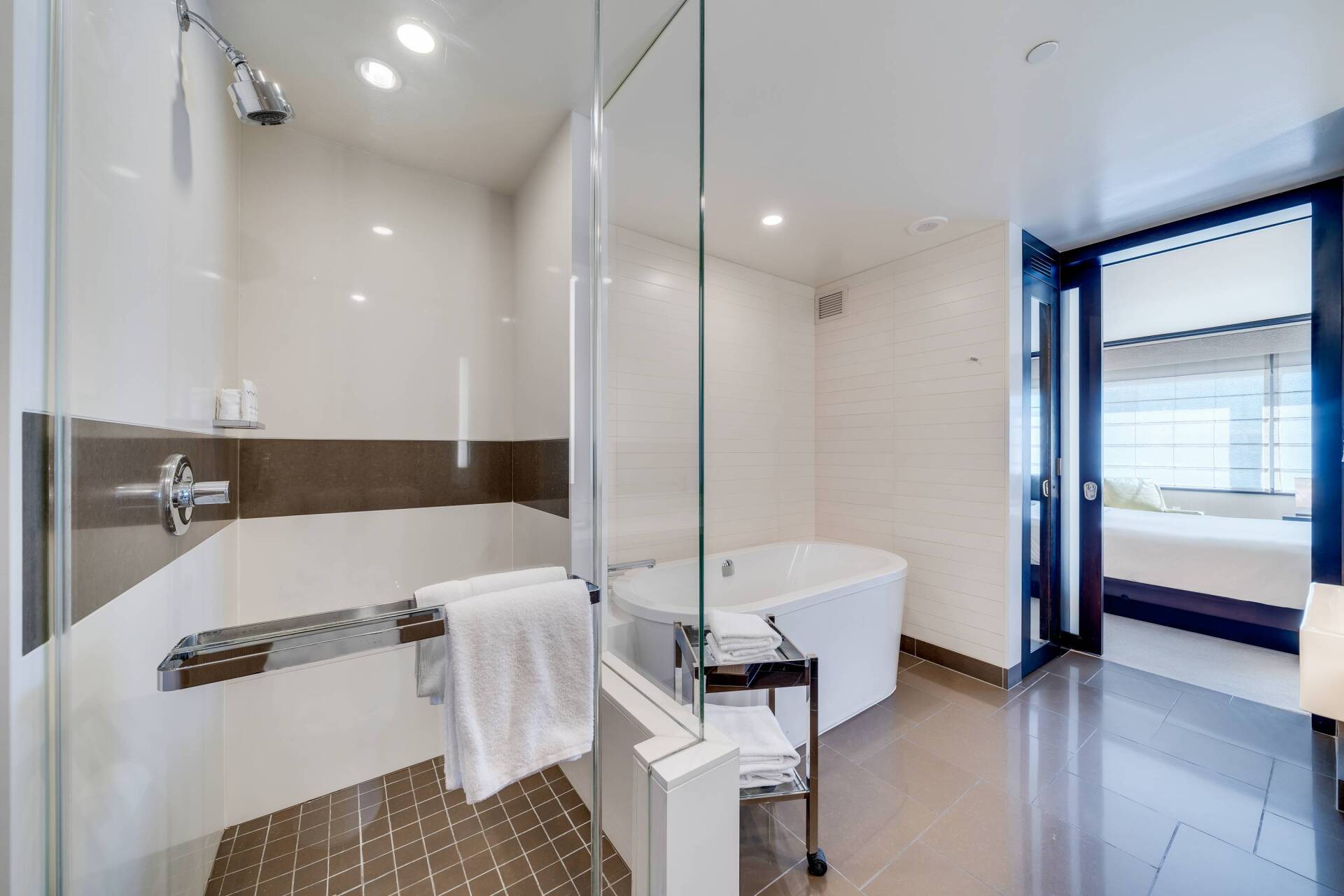 Modern bathroom with glass shower, freestanding tub, and open door to bedroom.