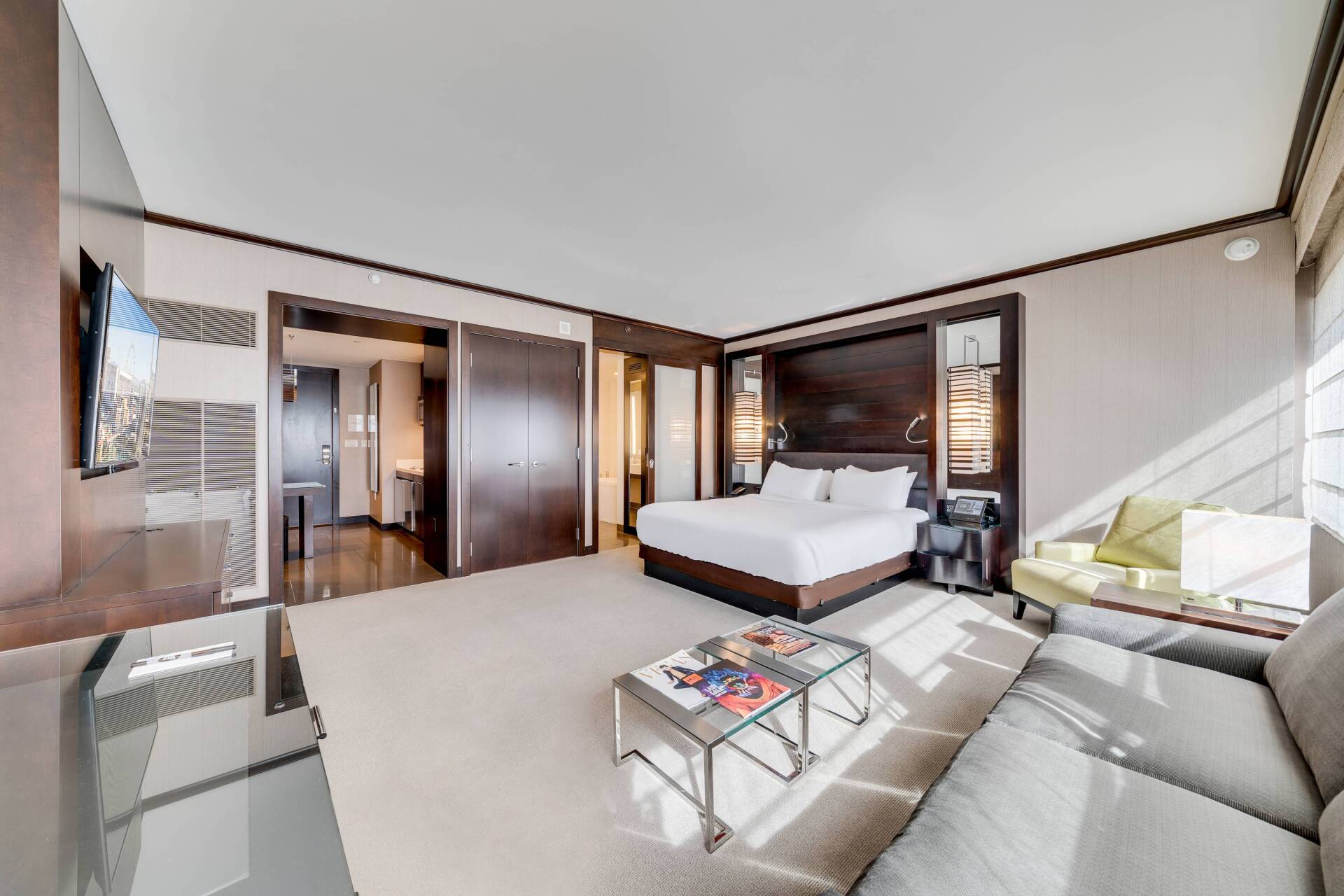 Modern hotel bedroom with king bed, sofa, coffee table, and wall-mounted TV in neutral tones