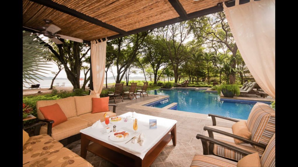 Luxury patio lounge with white table, cushioned seating, and a pool view under a thatched roof