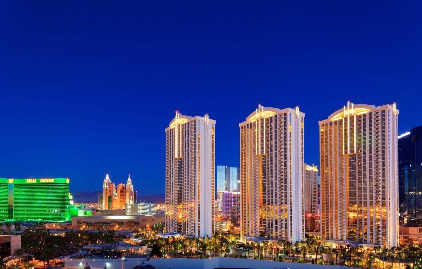City skyline at dusk with illuminated high-rise towers and a bright blue sky