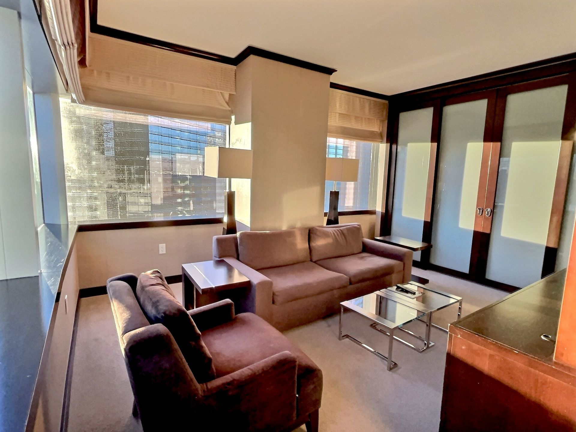 Bright living room with brown sofas, glass coffee table, and large windows overlooking city buildings