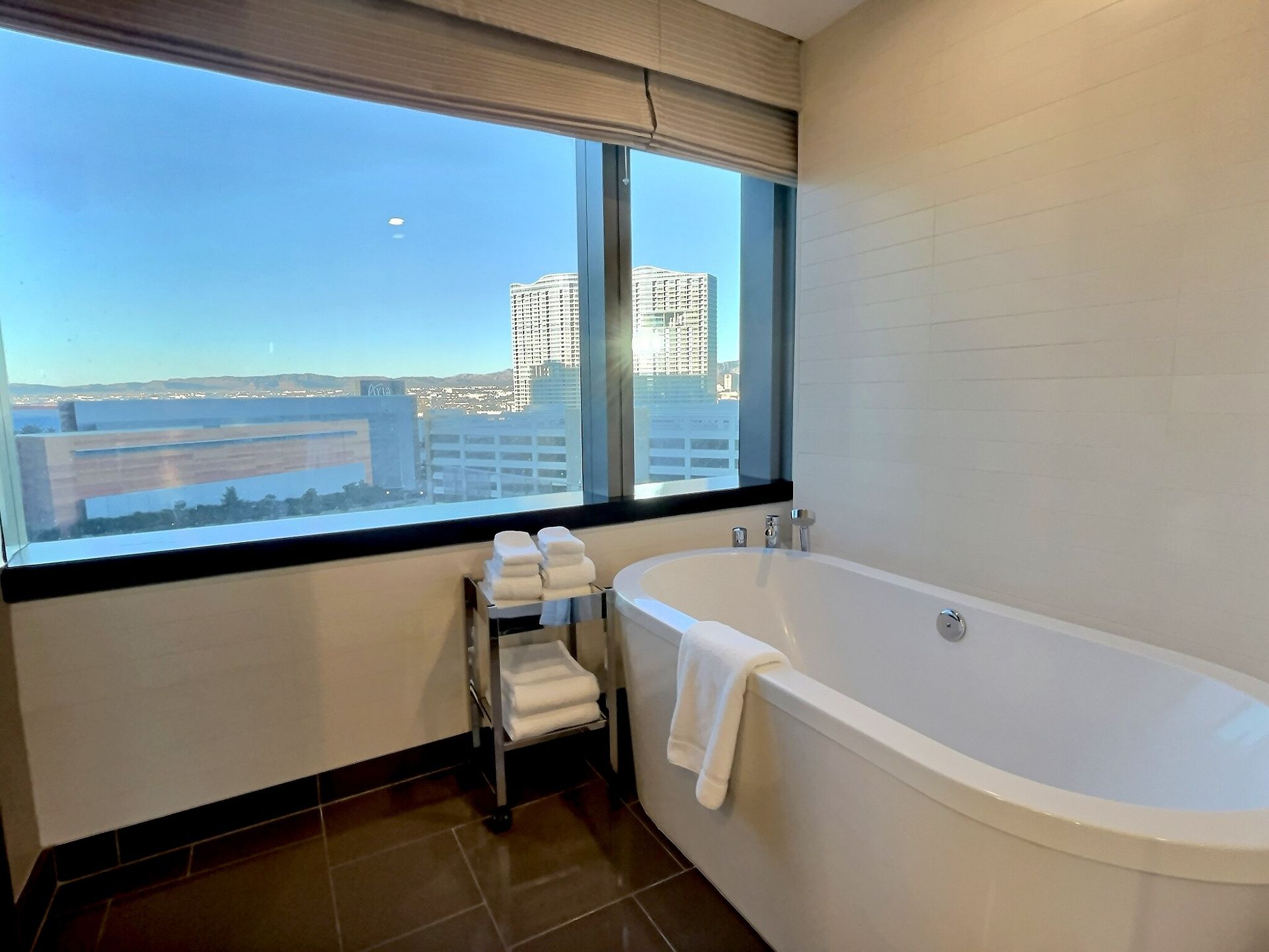 Modern bathroom with a large window, city view, and a white soaking tub with towels nearby