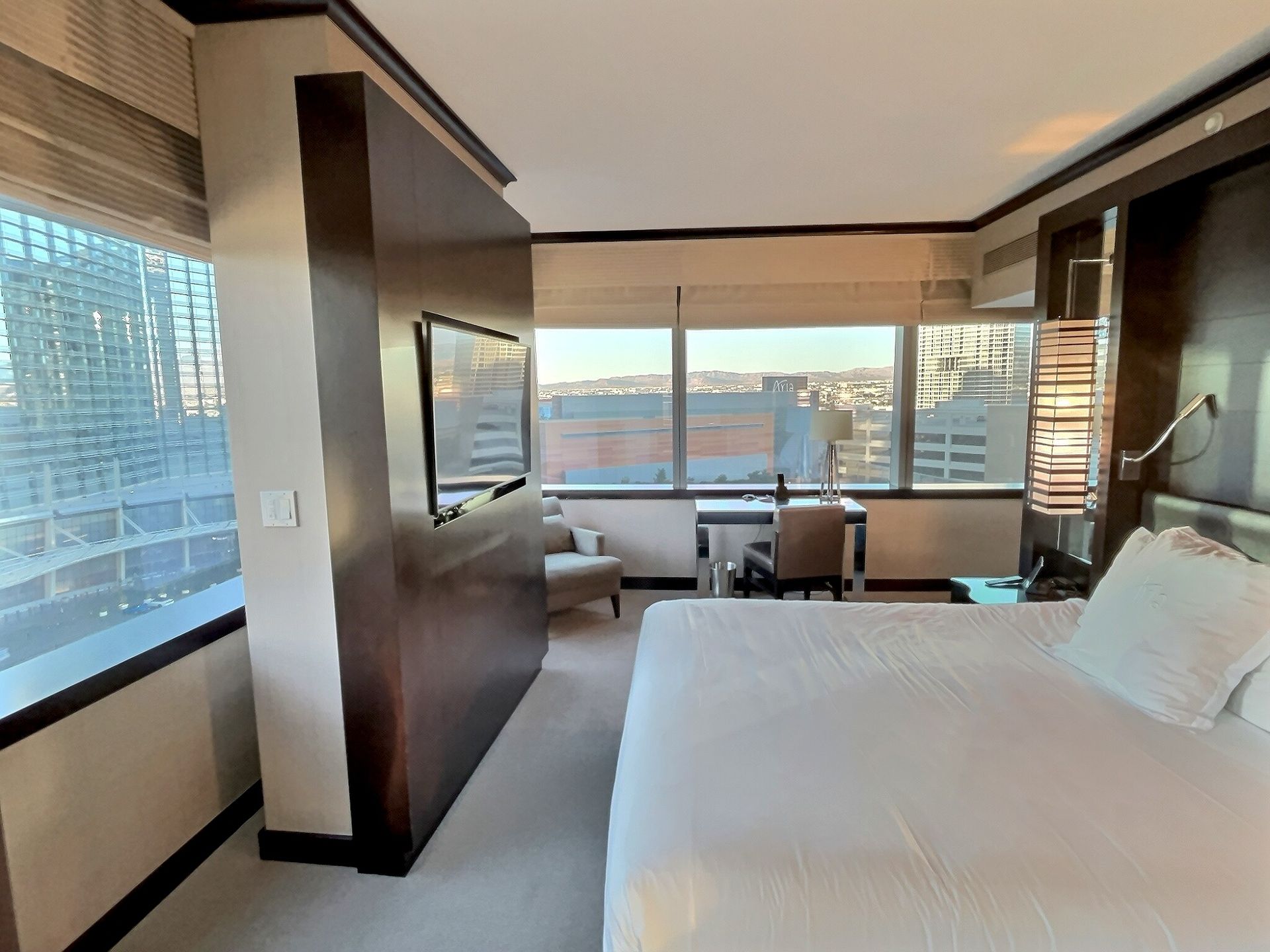 Modern hotel room with a bed, wall-mounted TV, desk, and floor-to-ceiling windows with city view
