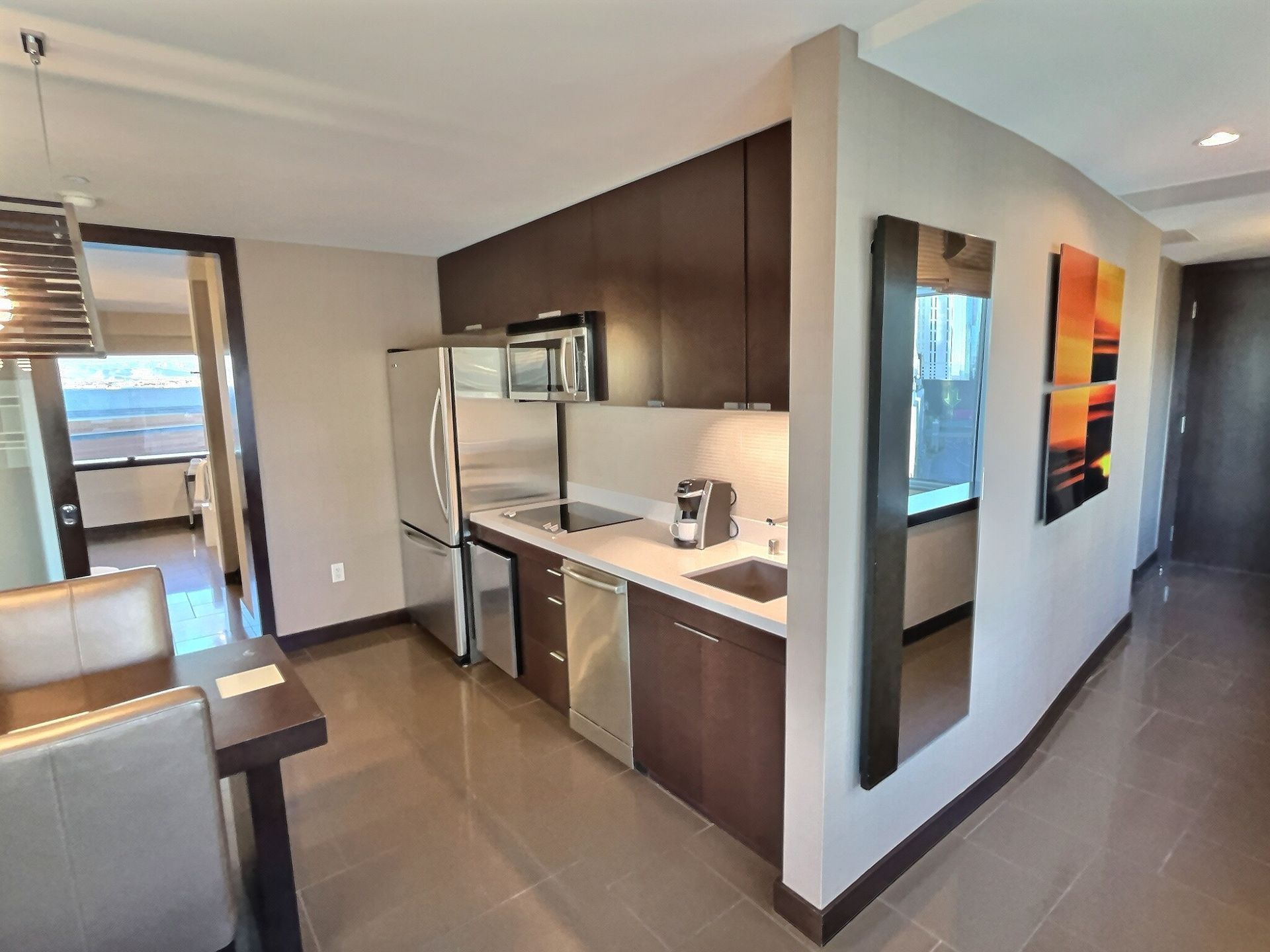 Modern apartment kitchen with dark wood cabinets, stainless appliances, and a narrow hallway to a bright living area