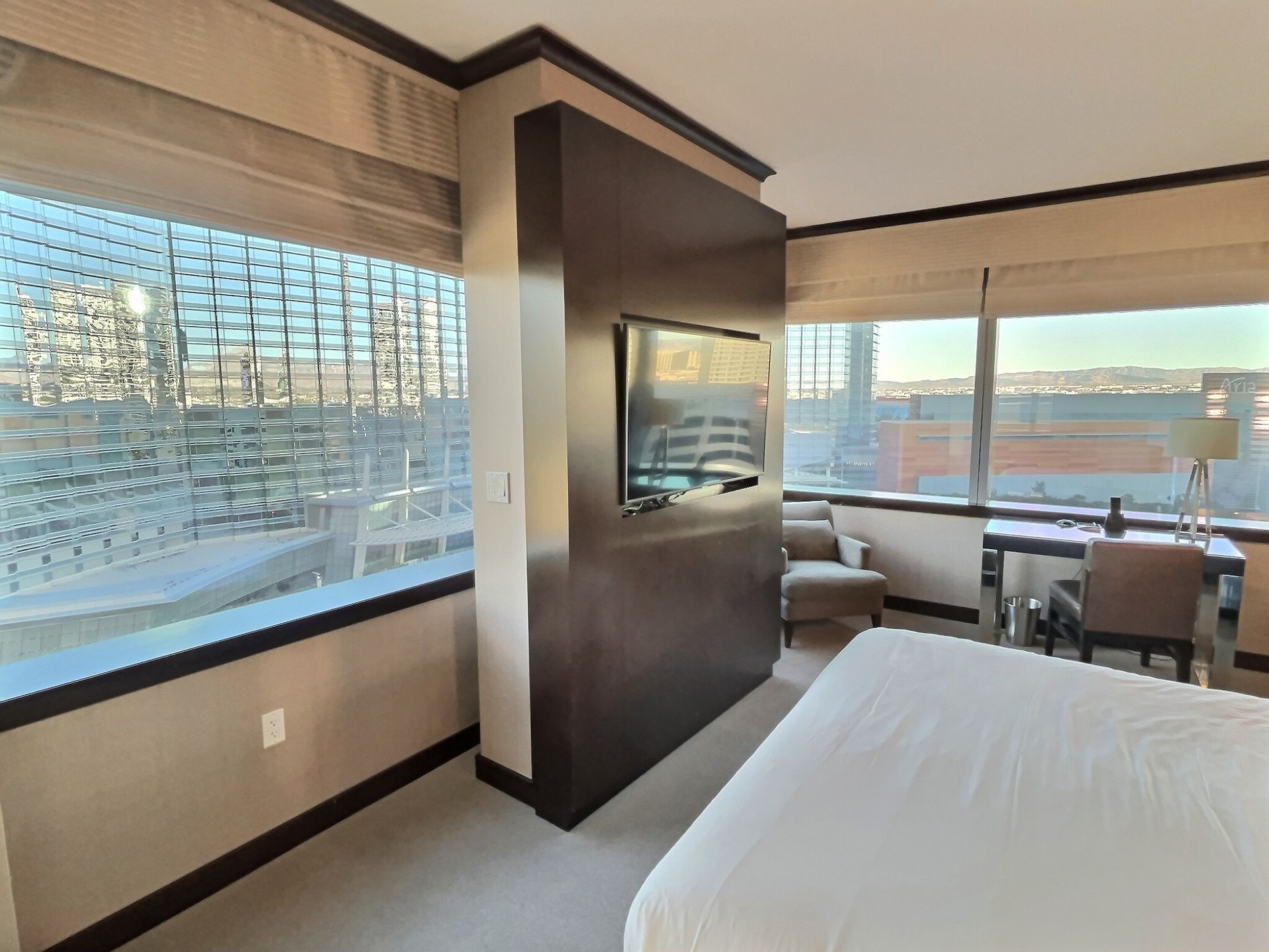 Hotel room with two windows, a bed, chairs, and a wall-mounted TV overlooking a city view