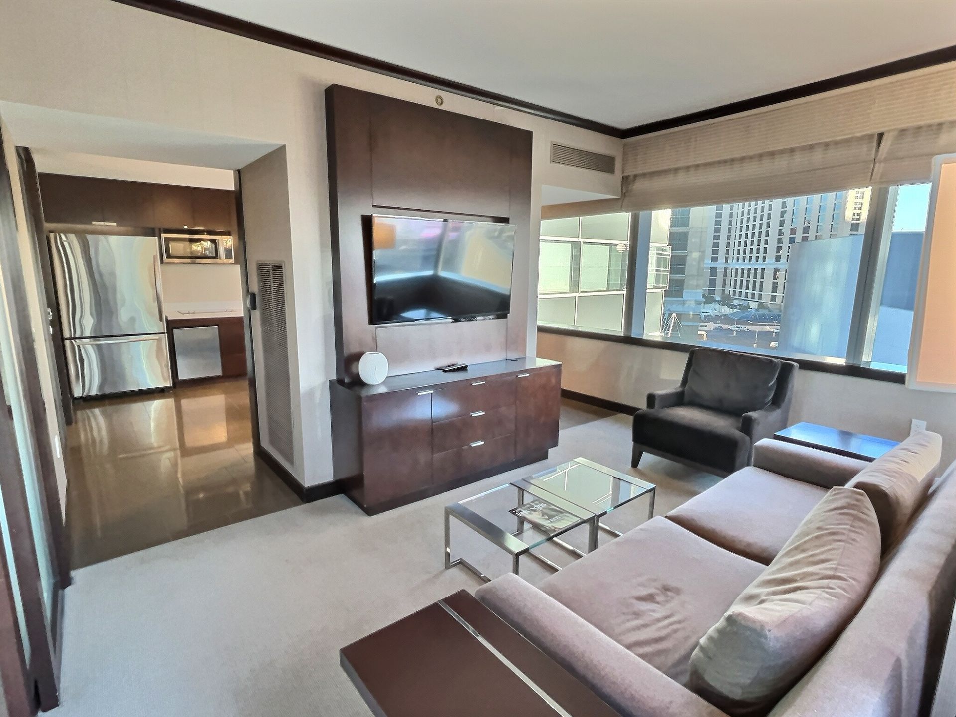 Modern living room with sofa, TV, glass coffee table, and large windows overlooking city buildings