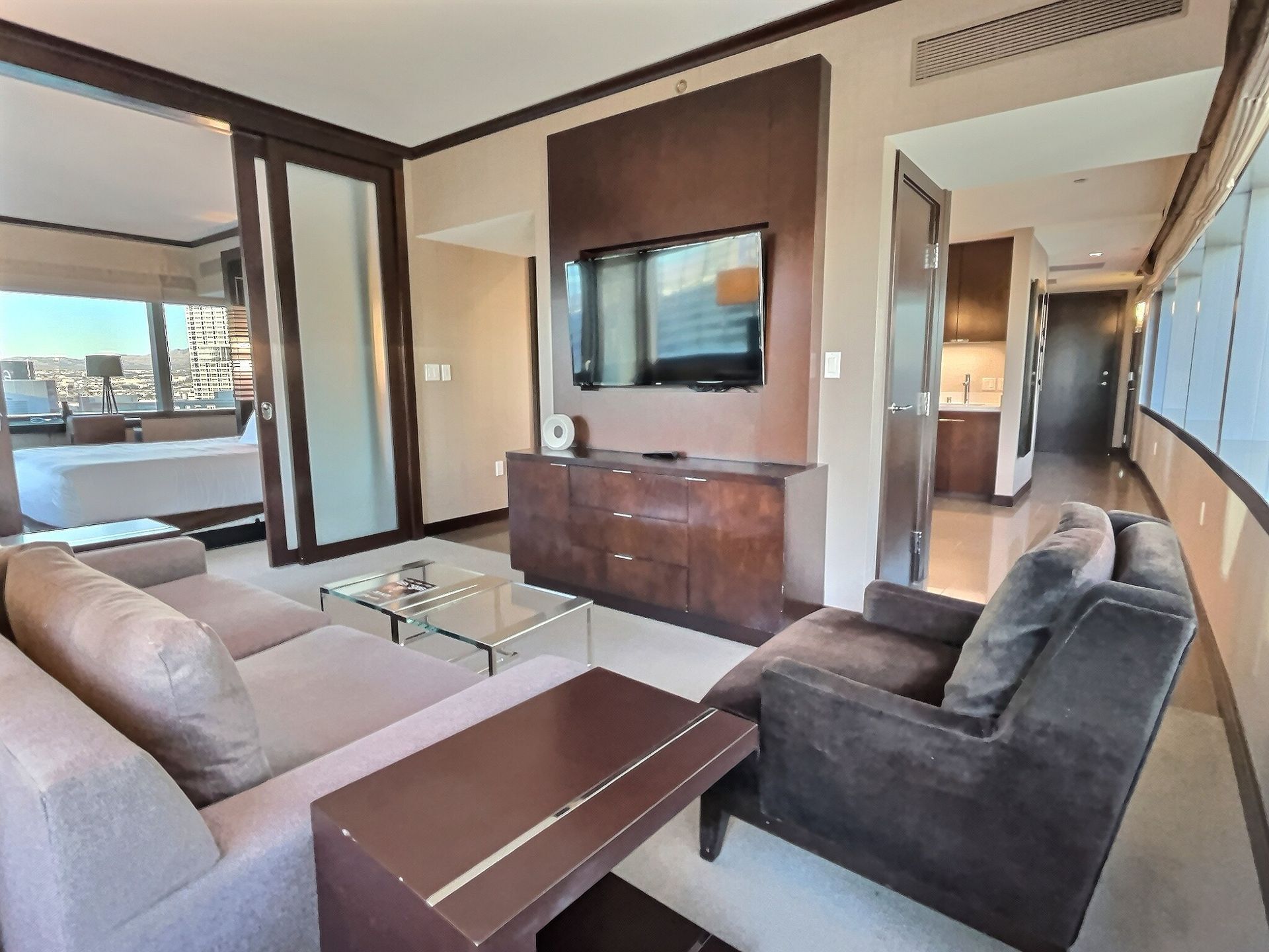 Modern hotel suite living area with gray sofas, coffee table, wall-mounted TV, and city view through sliding doors