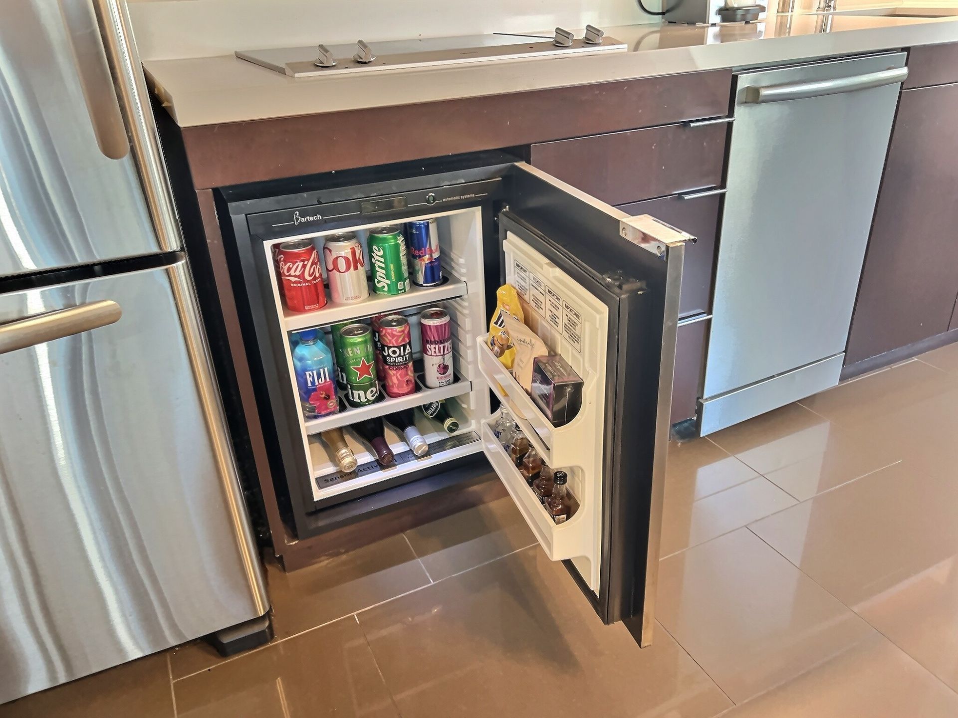Open mini fridge in a kitchen, stocked with sodas and drinks.