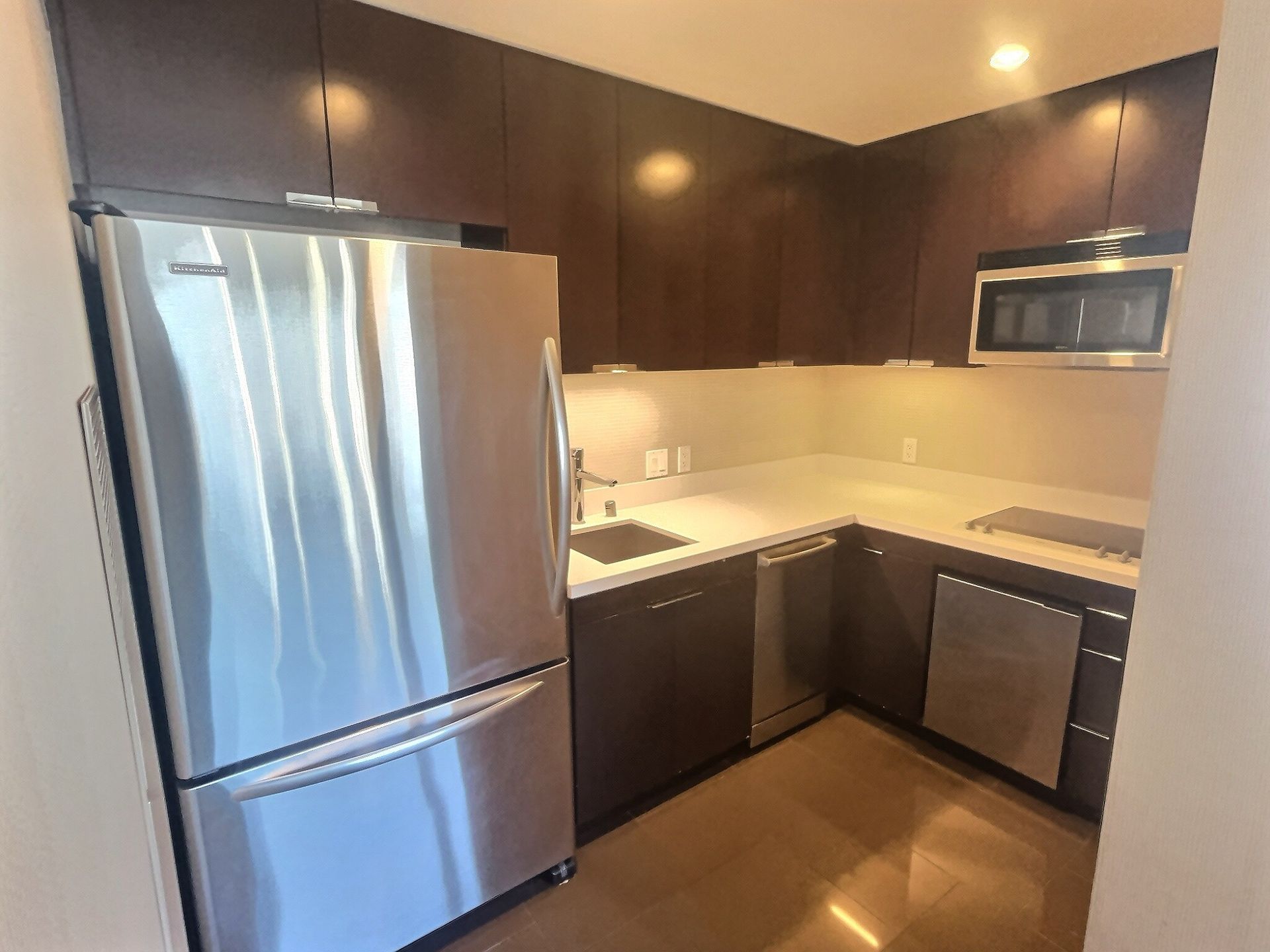 Modern kitchen with stainless steel fridge, dark cabinets, and a corner sink beneath a microwave.