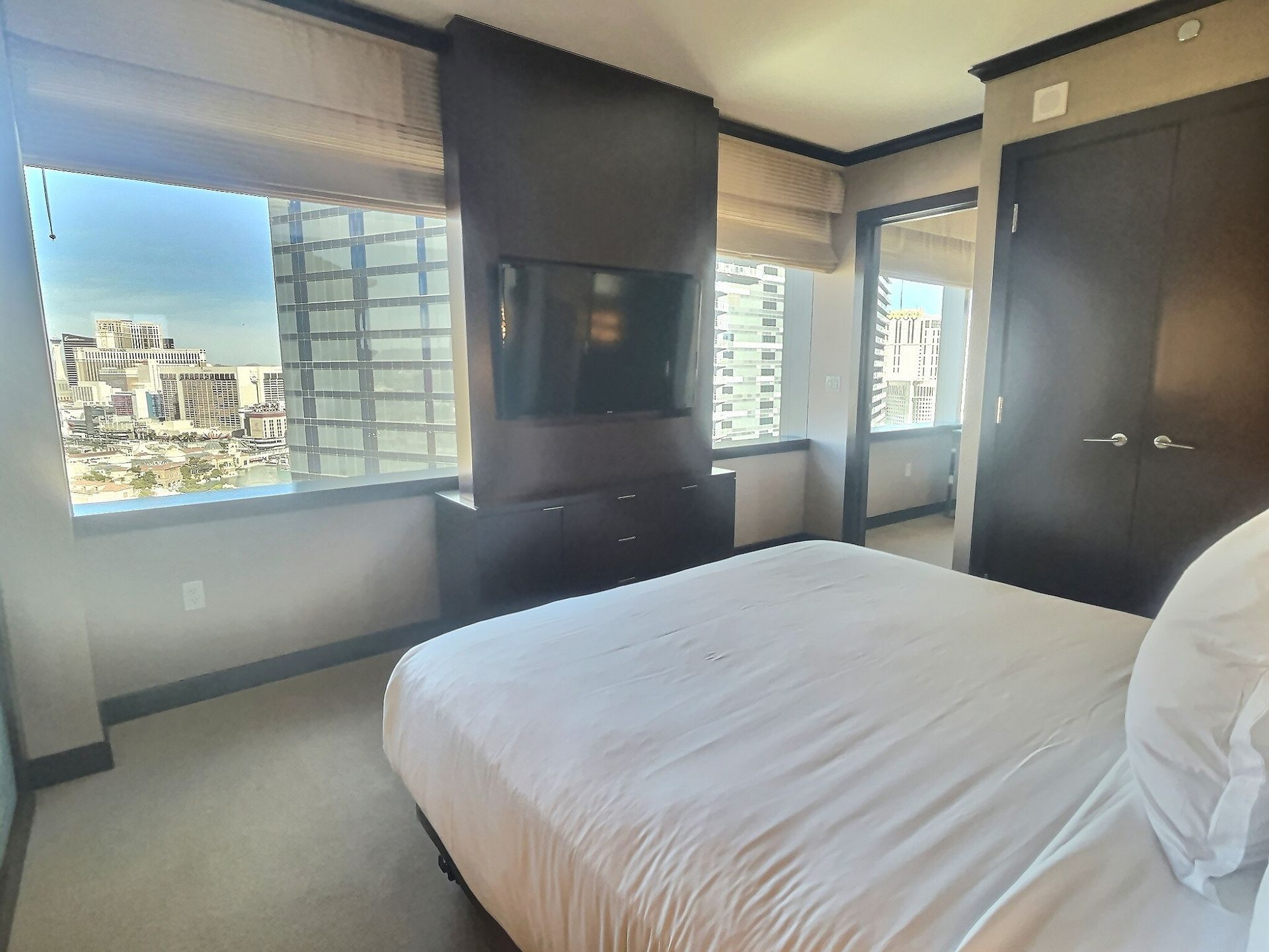 Bright bedroom with large windows, white bed, dark wood cabinets, and city view