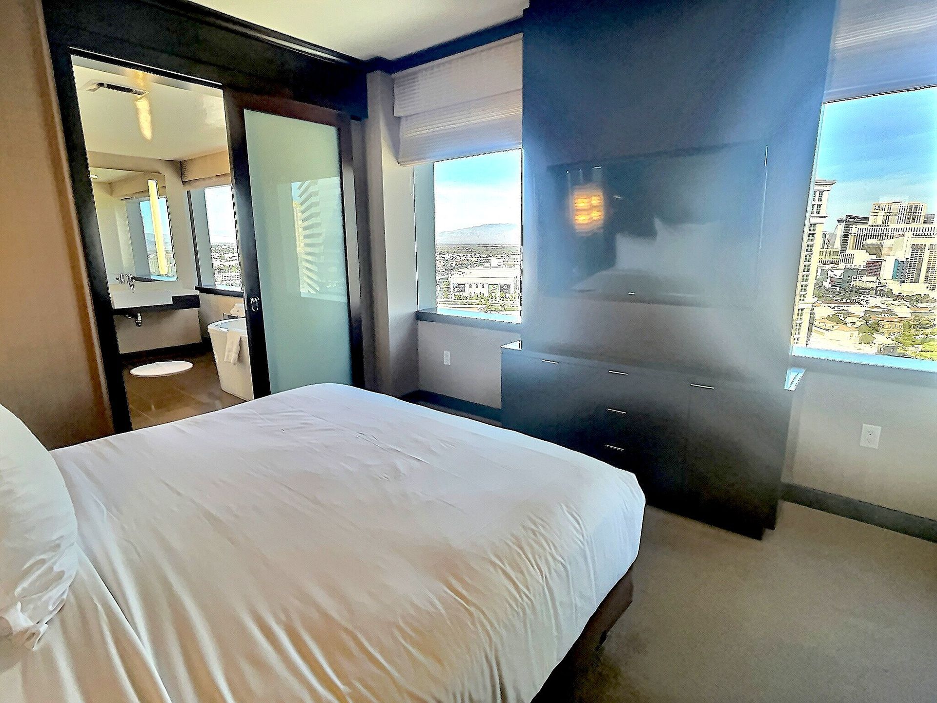 Modern bedroom with white bed, dark wood accents, and large windows overlooking a city view