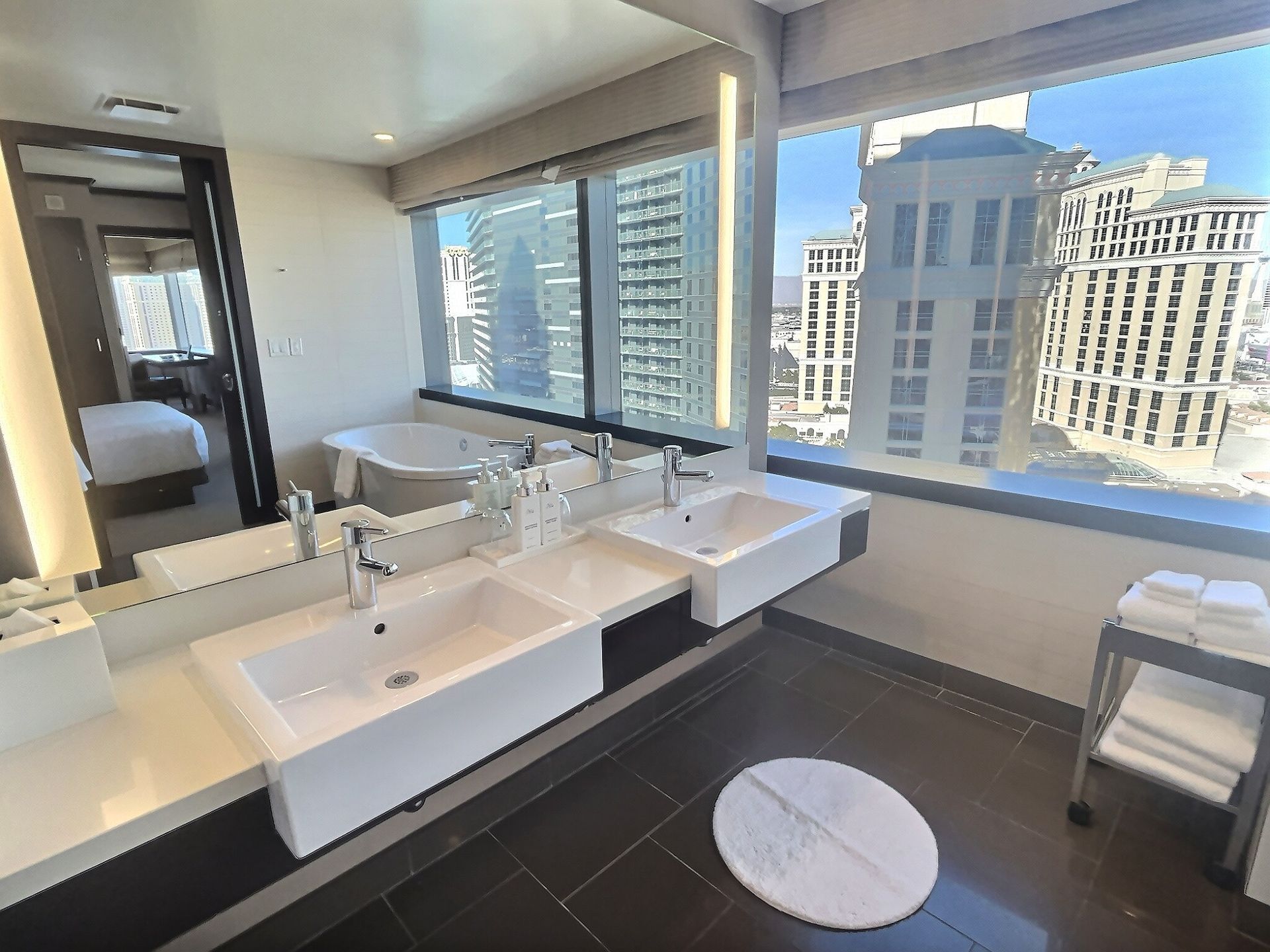 Modern bathroom with double sinks, large mirror, city-view windows, and a round floor mat
