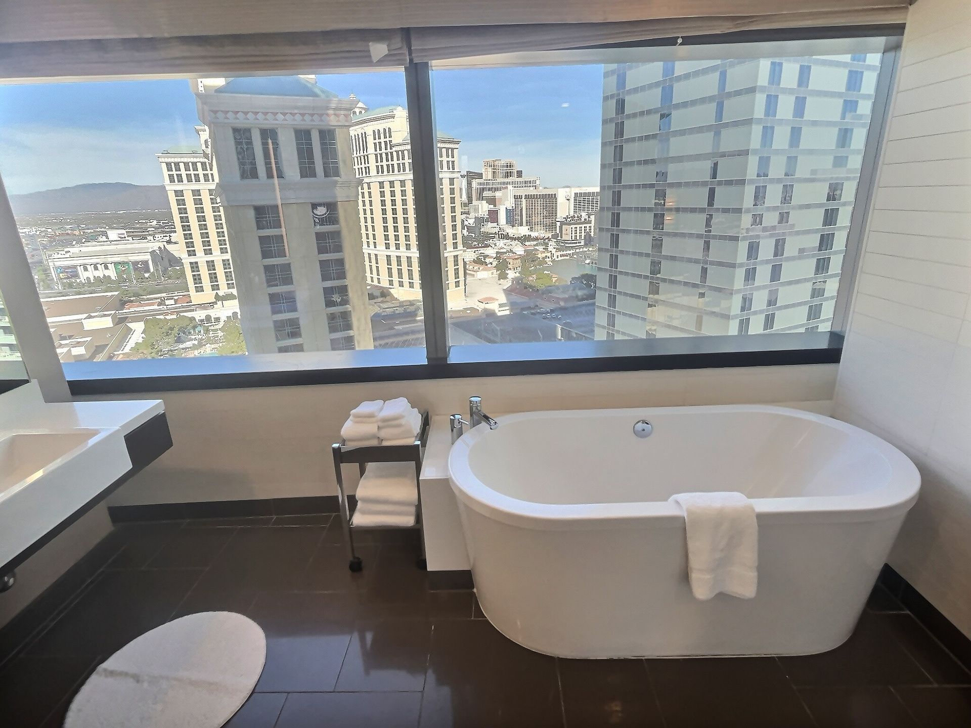Modern bathroom with a white bathtub, dark floor, and large window overlooking city buildings