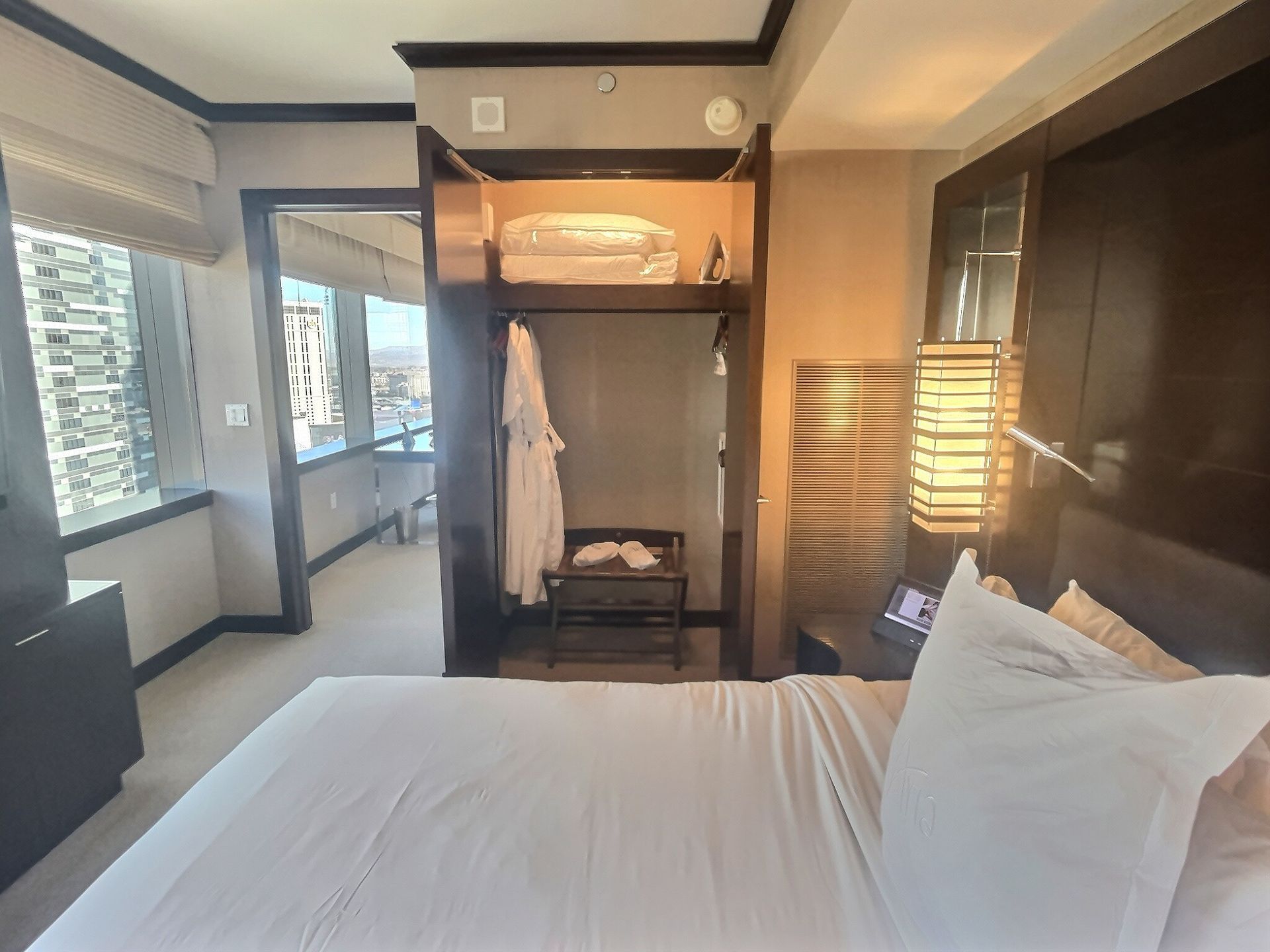 Modern hotel bedroom with white bed, wooden wardrobe, and large windows with blinds