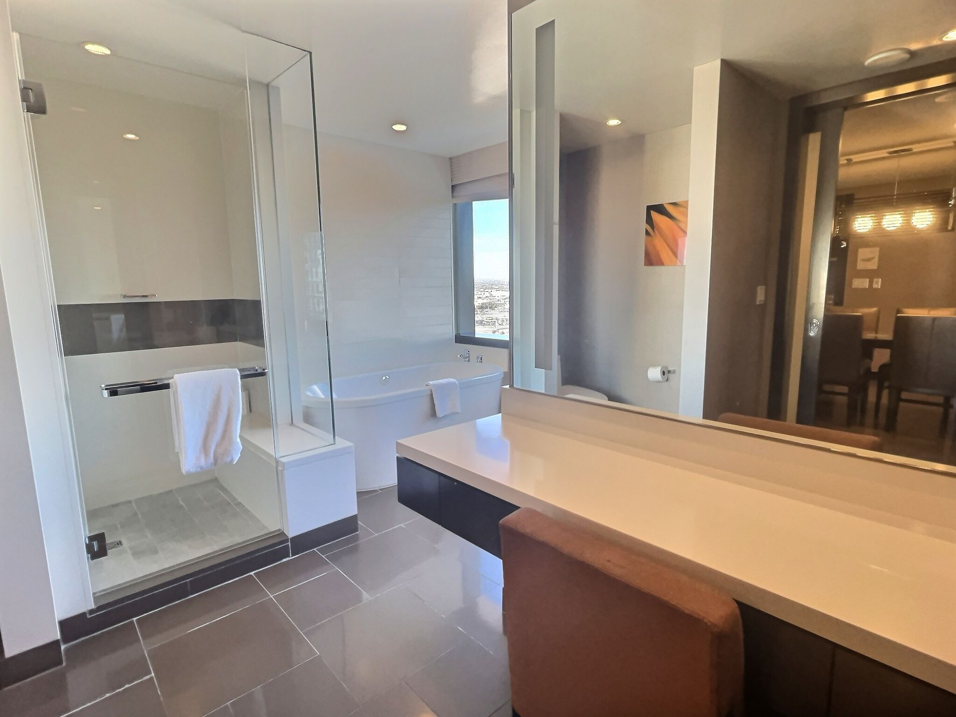 Modern bathroom with glass shower, large mirror, and vanity with beige countertop and brown stool.