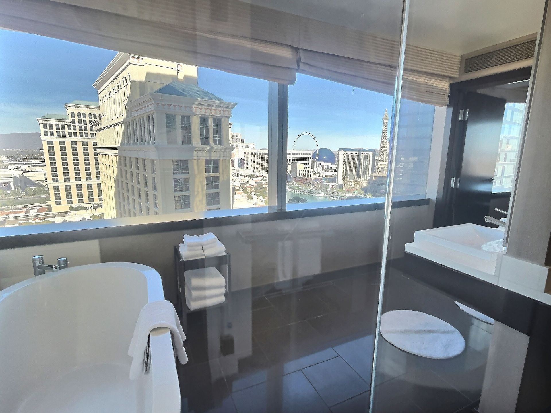 Modern bathroom with a large window overlooking city buildings and ocean, white tub and sink in view