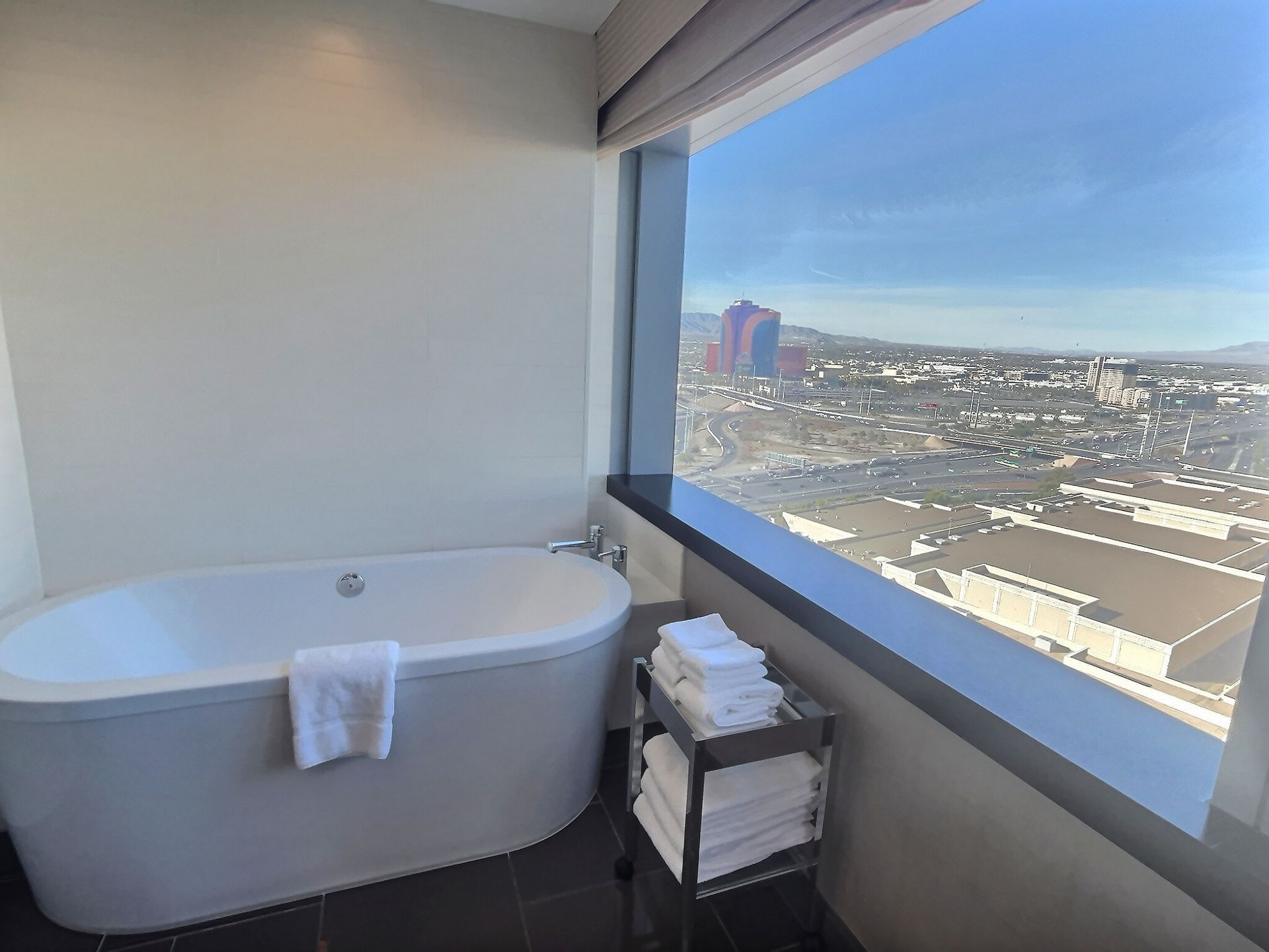 Modern bathroom with a freestanding tub, towel stack, and city view through a large window