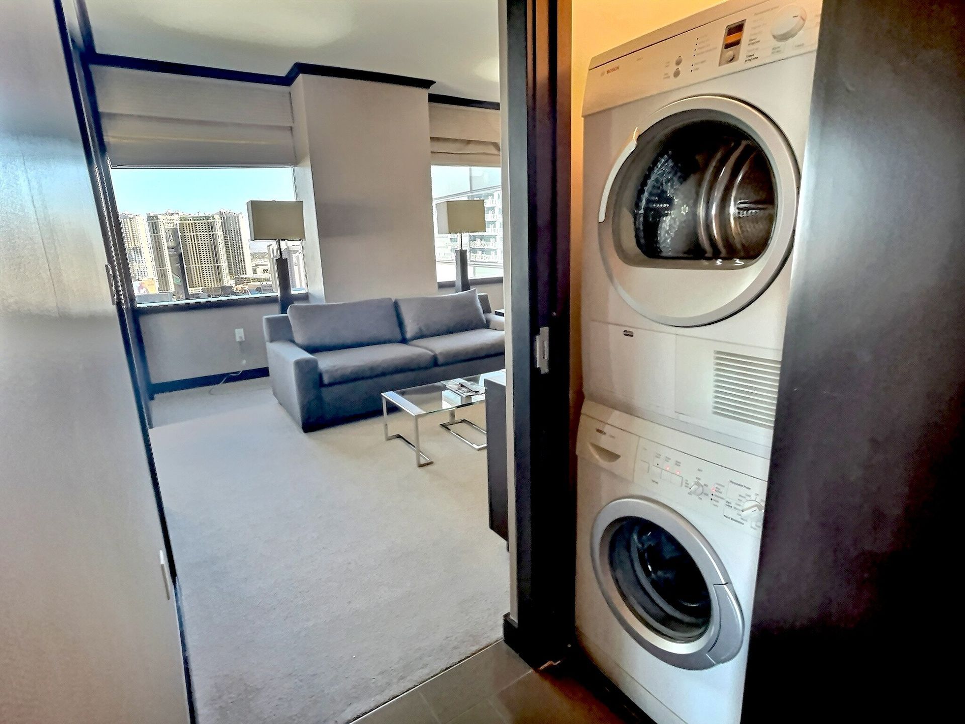 Stacked washer and dryer in a doorway, with a bright living room and gray sofa beyond.
