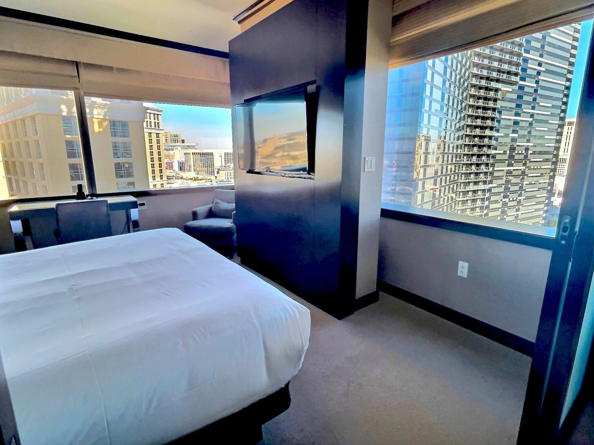 Modern hotel room with a bed, TV, and large windows overlooking tall city buildings